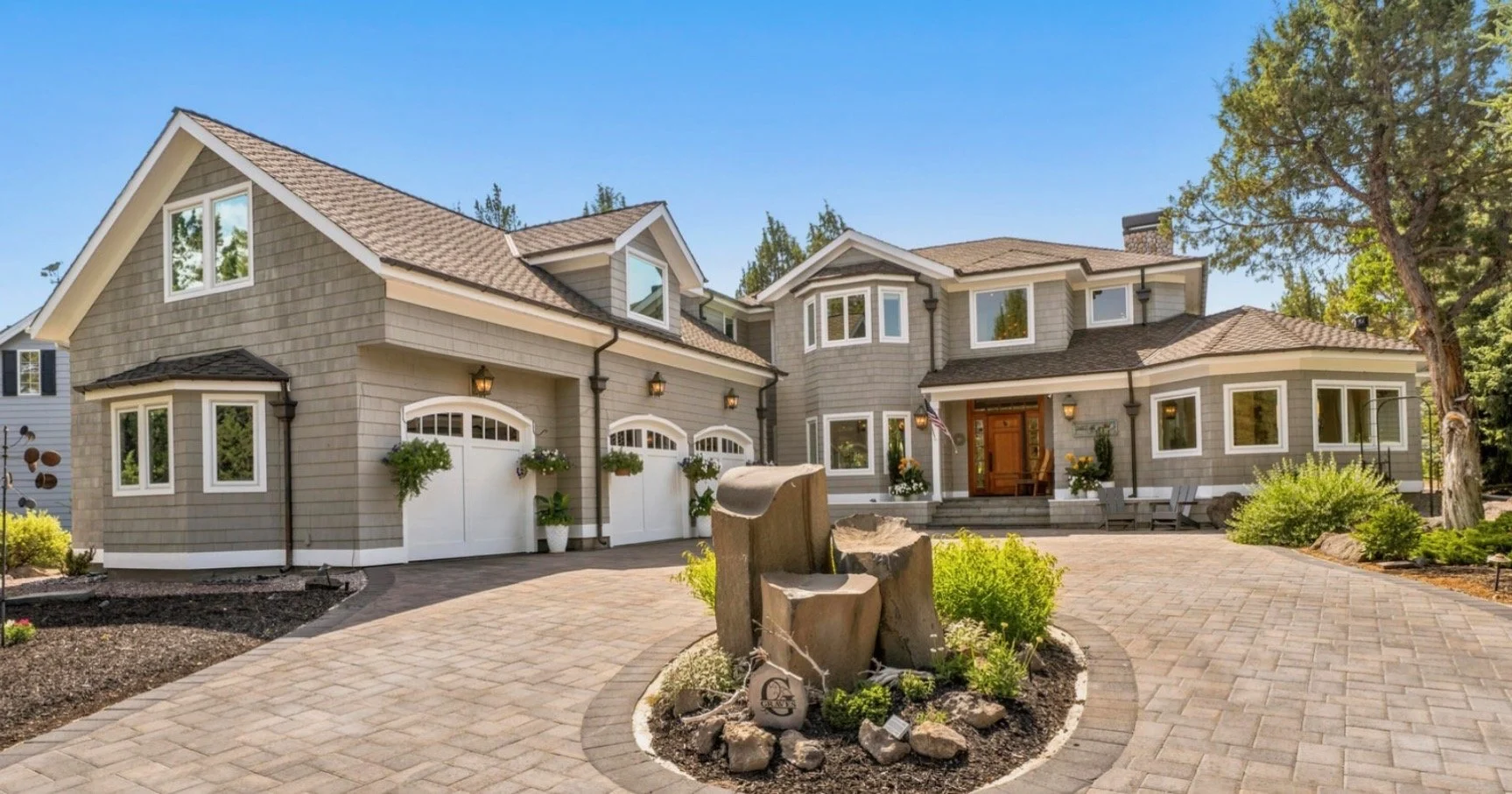 A large, modern suburban house with gray siding and a landscaped front yard. The house features multiple windows, a wooden front door, and a driveway with a rock centerpiece.