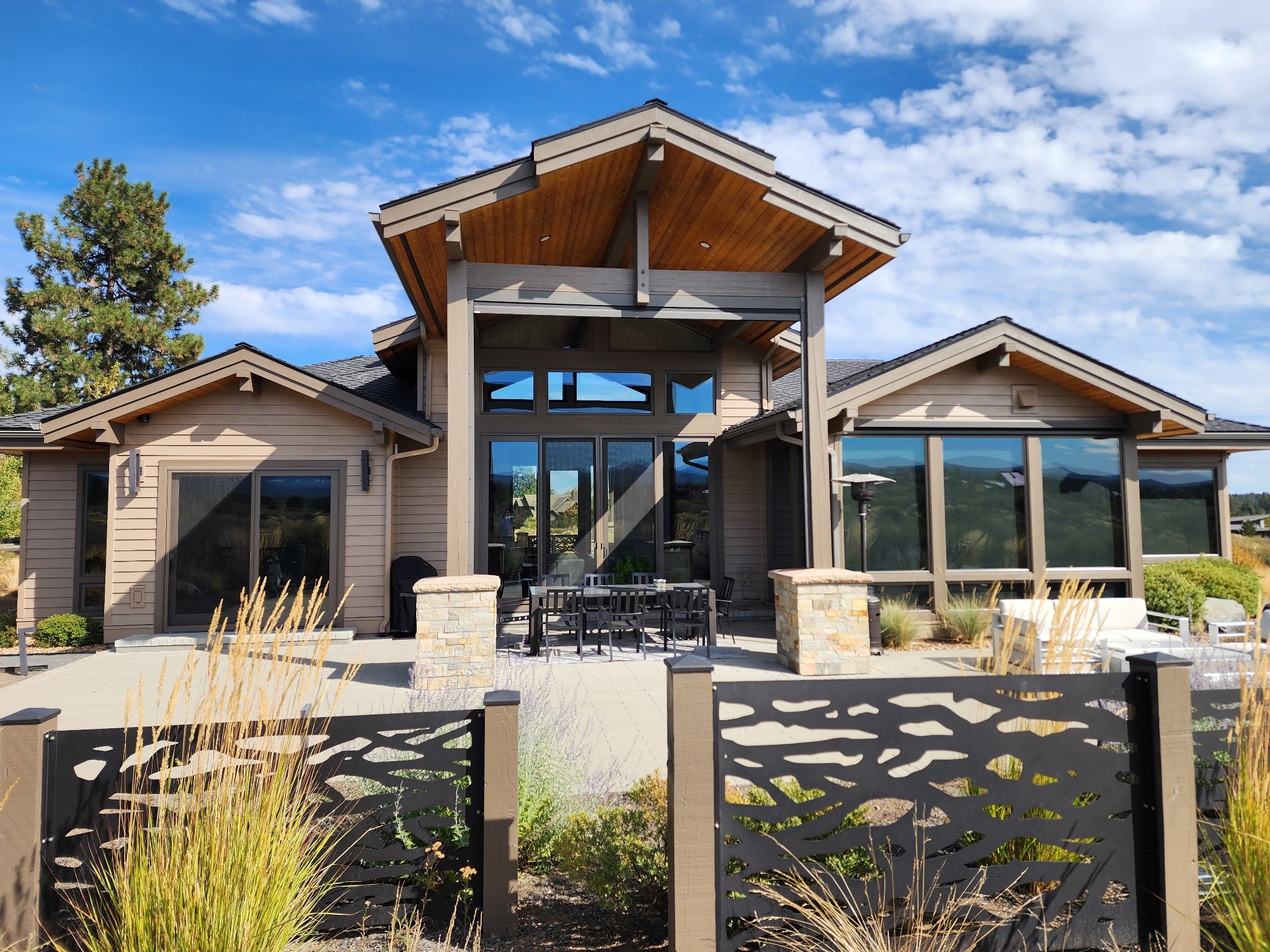 Modern house with large windows and a backyard patio, surrounded by a decorative black fence and drought-tolerant plants, under a bright blue sky with scattered clouds.