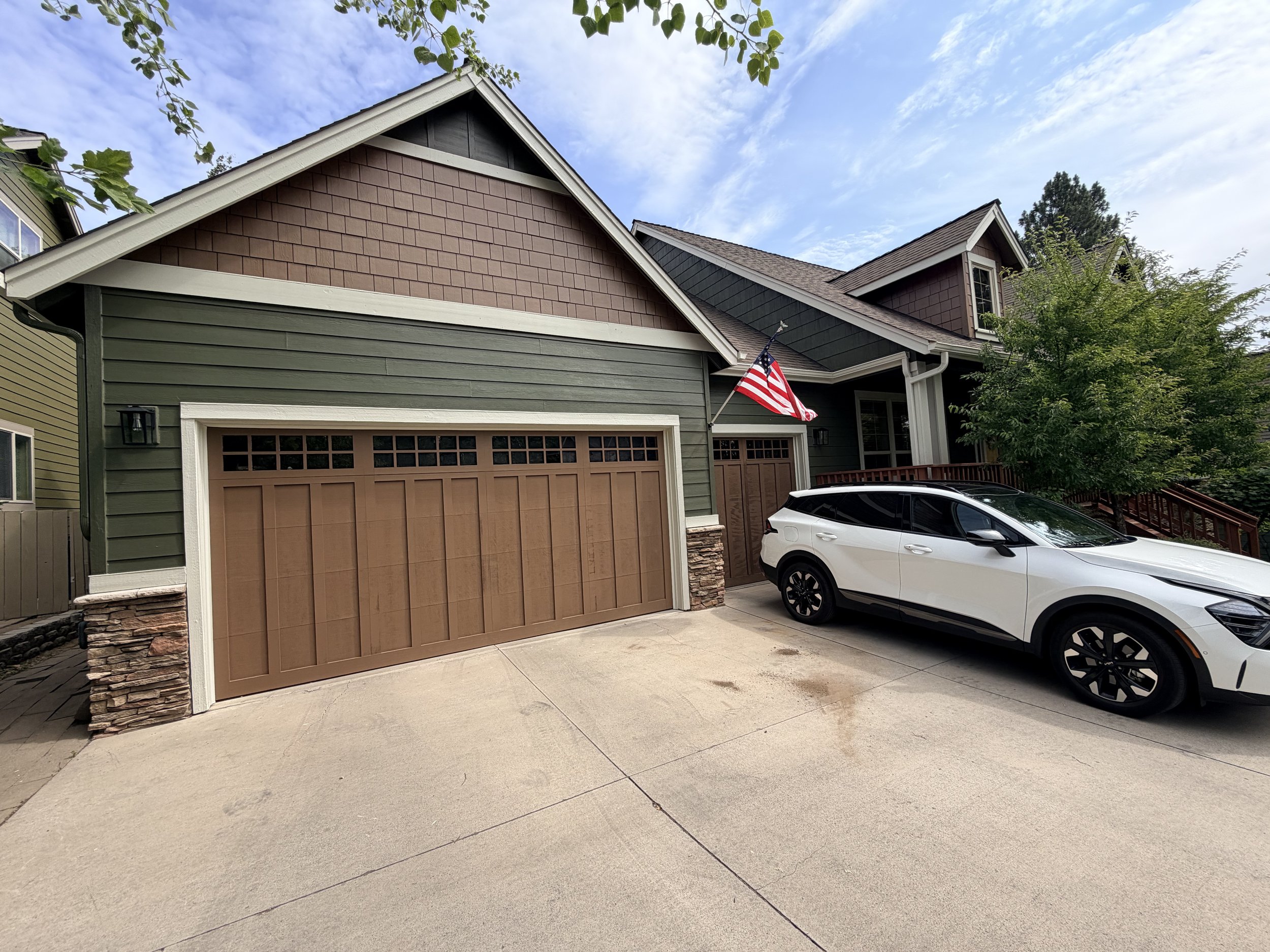 A two-story house with a green exterior, brown garage door, and brown shingle siding on the upper part. An American flag is mounted on the front of the house. A white car is parked in the driveway, and there are trees and a blue sky with clouds in th
