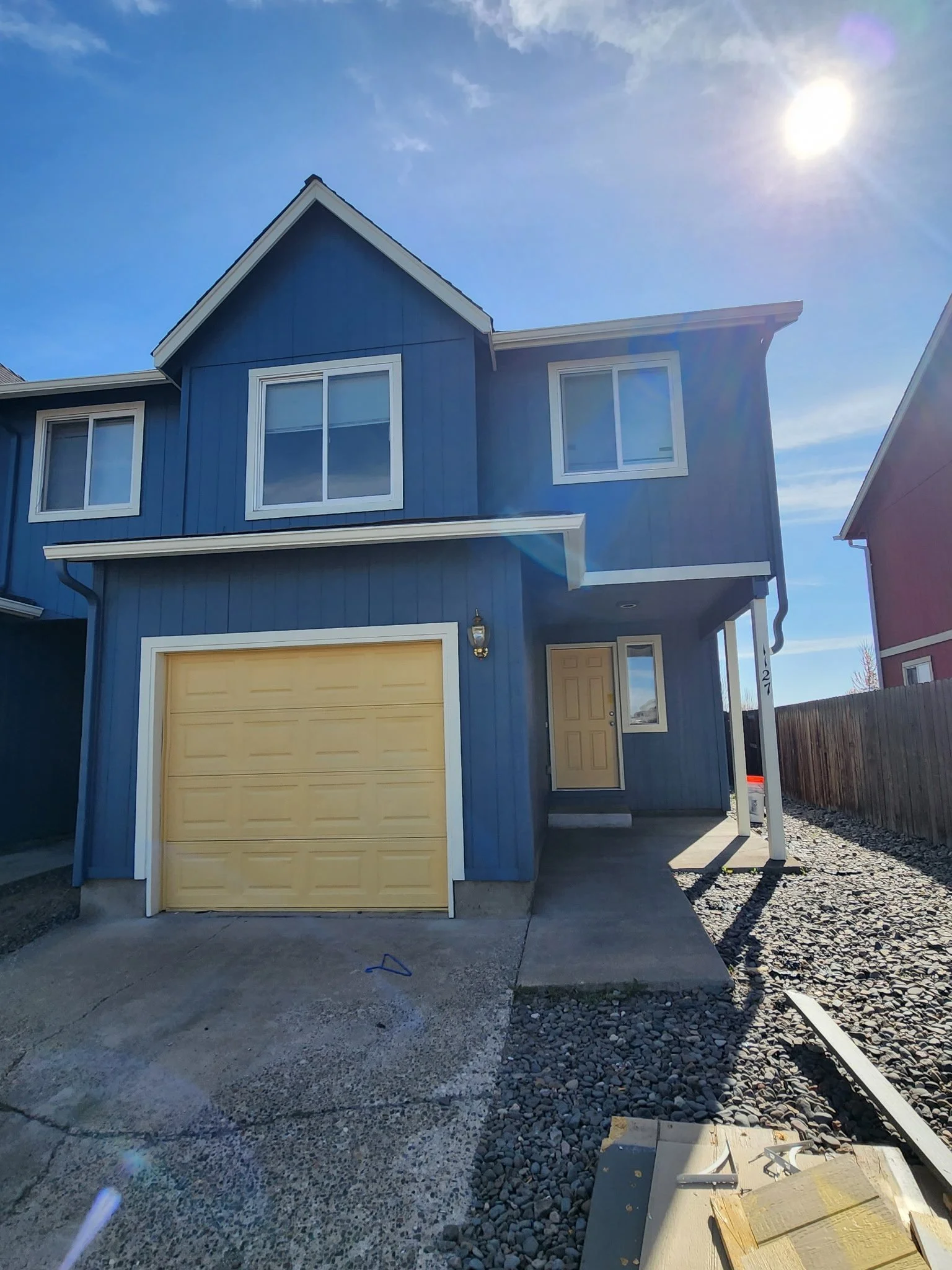Front of a two-story blue house with white trim. The house has a yellow garage door, a front door, and two windows on the second floor. The driveway is under construction with gravel and building materials visible, and the sky is clear with the sun s