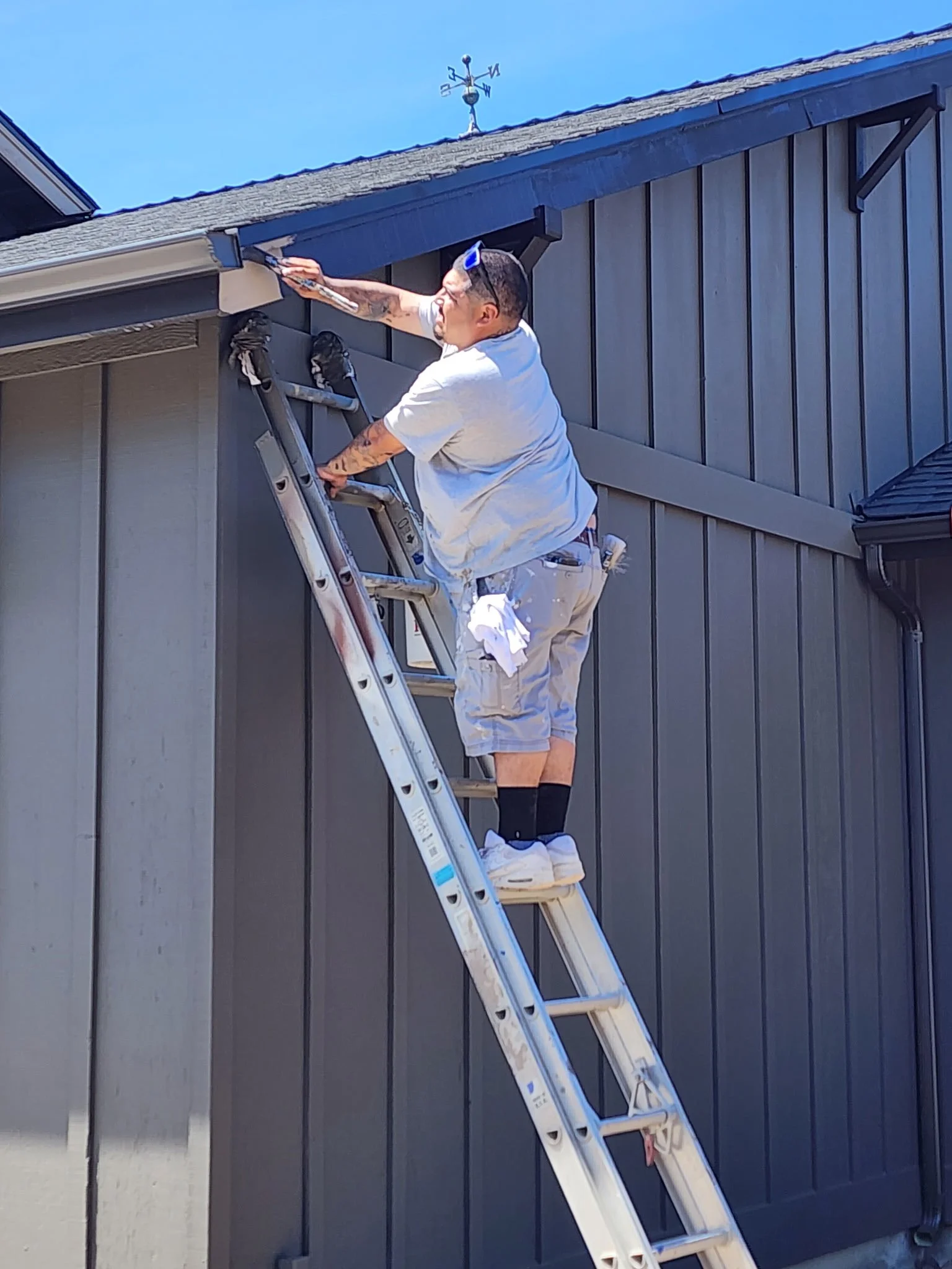 A man standing on a ladder cleaning the gutters of a house with black siding and a shingled roof under a clear blue sky.