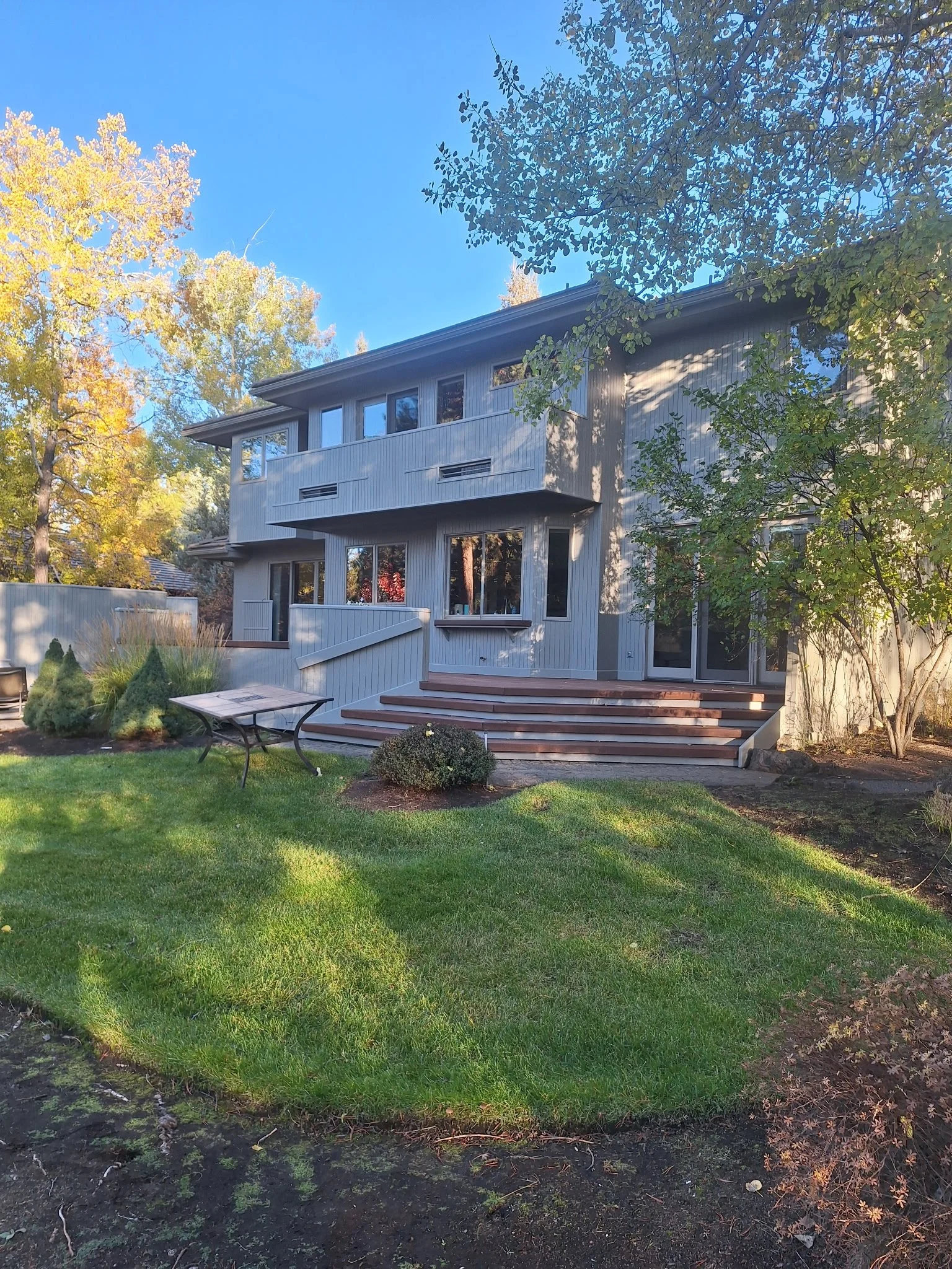 A modern, multi-story house with large windows and a light-colored exterior, surrounded by a manicured lawn and trees with autumn foliage, under a clear blue sky.