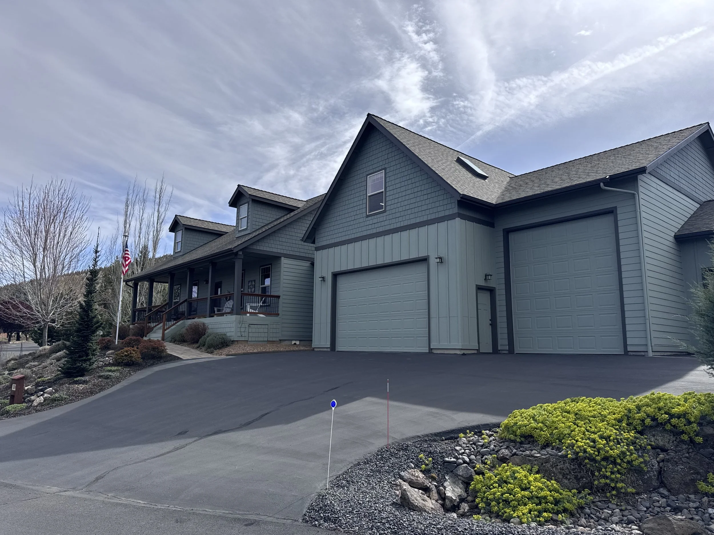 A modern gray house with a gable roof, attached garage, and a landscaped driveway. The sky is partly cloudy, with leafless trees and a few evergreen trees in the background, and an American flag near the house.