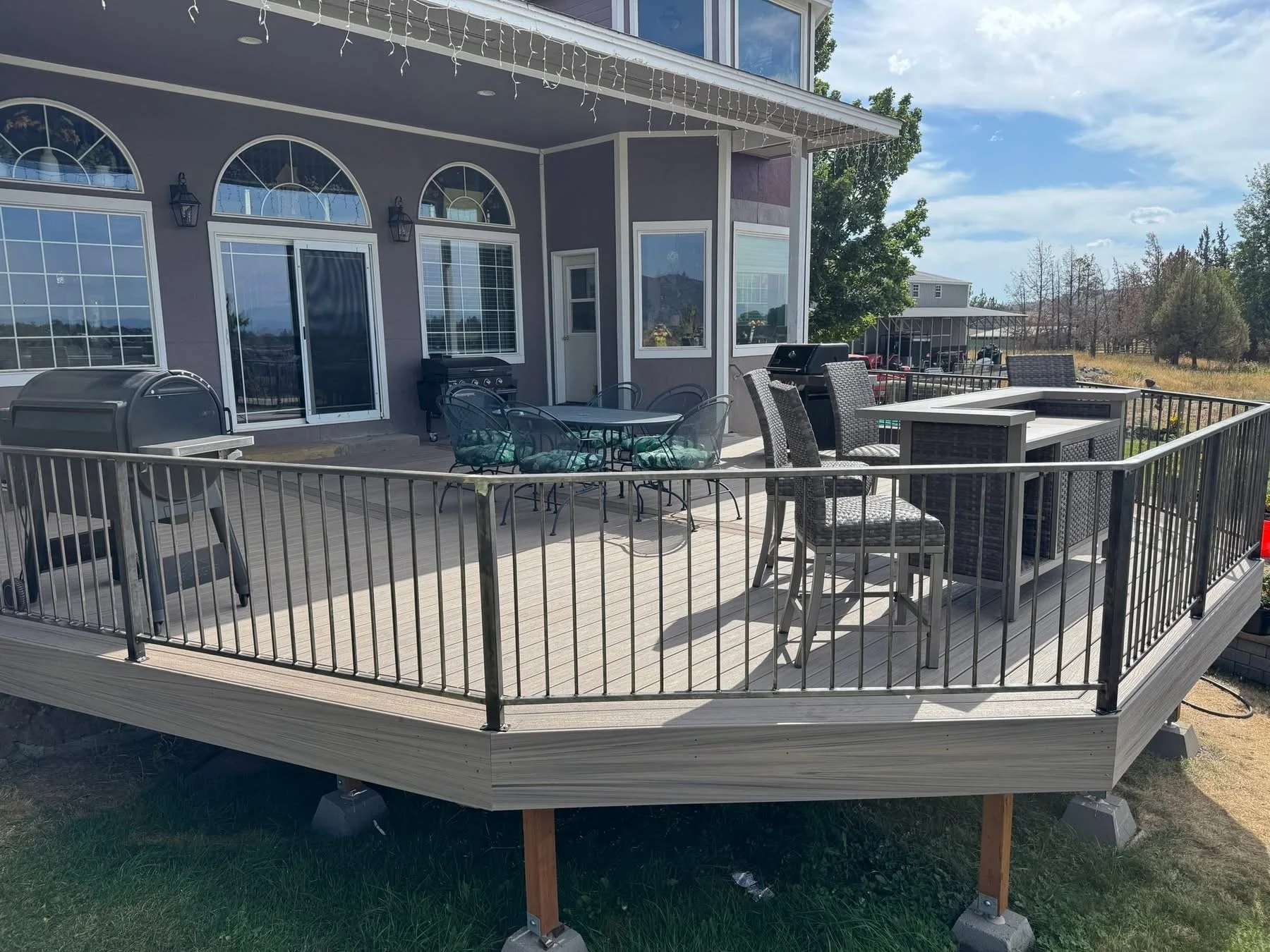 A large outdoor wooden deck attached to a house with gray siding, fenced with black metal railing. The deck has patio furniture including a round table with four chairs, a bar-height table with two chairs, and a grill, with a grassy lawn underneath.
