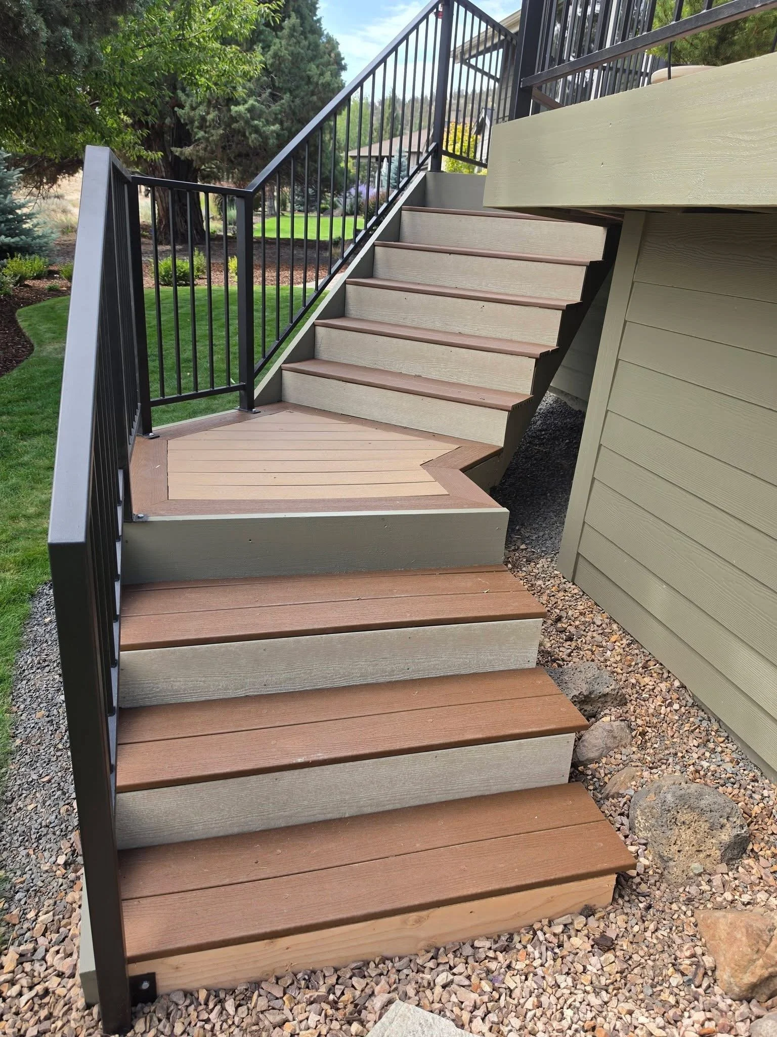 Outdoor staircase with wooden steps and black metal railing, leading up to a deck in a landscaped yard with trees and grass.