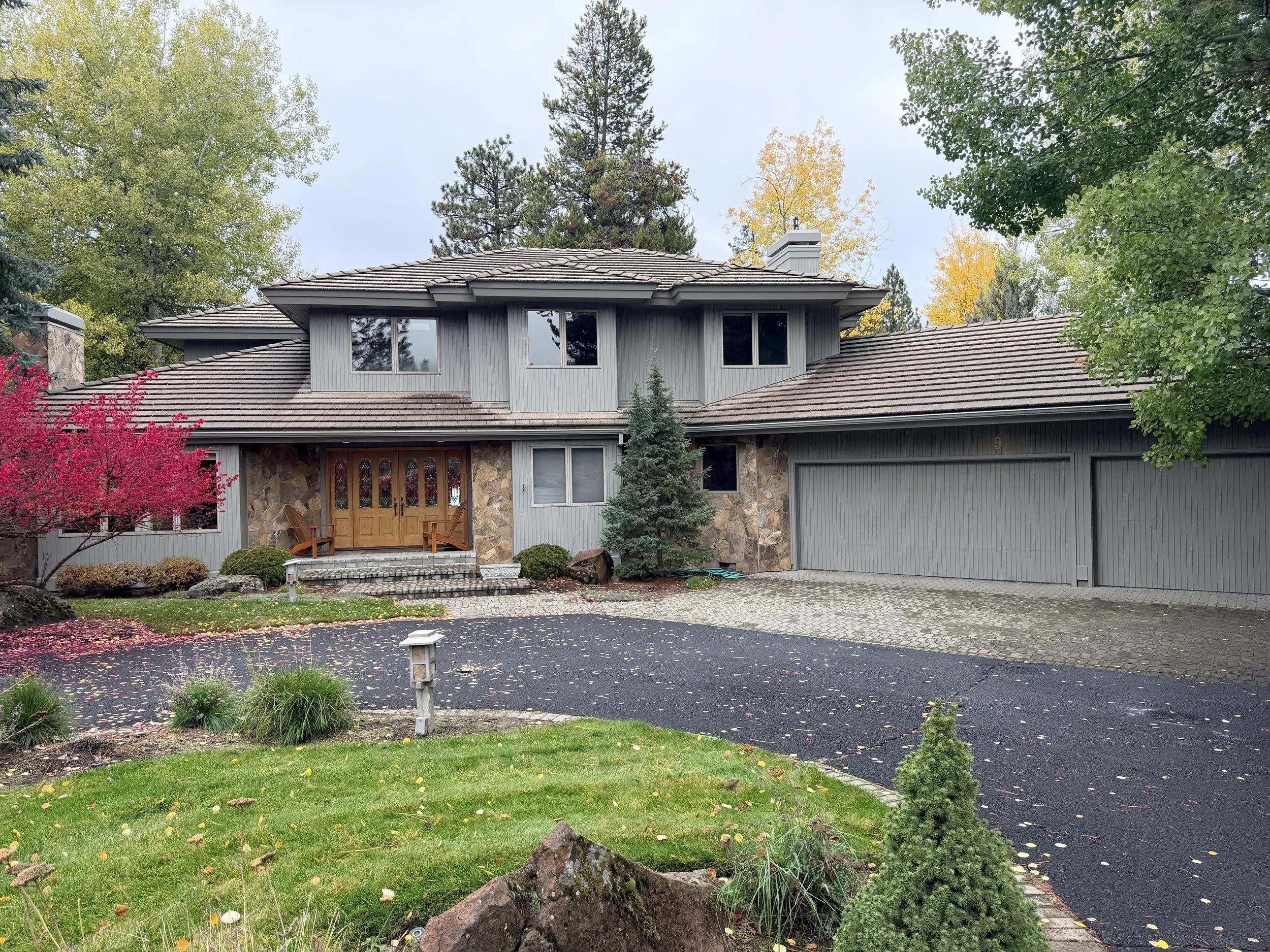 A two-story house with gray exterior, stone accents, and a brown tiled roof. The front yard has green grass, a pink flowering tree, and various shrubs. The driveway is paved and leads to a closed garage. Tall trees with autumn foliage are in the back