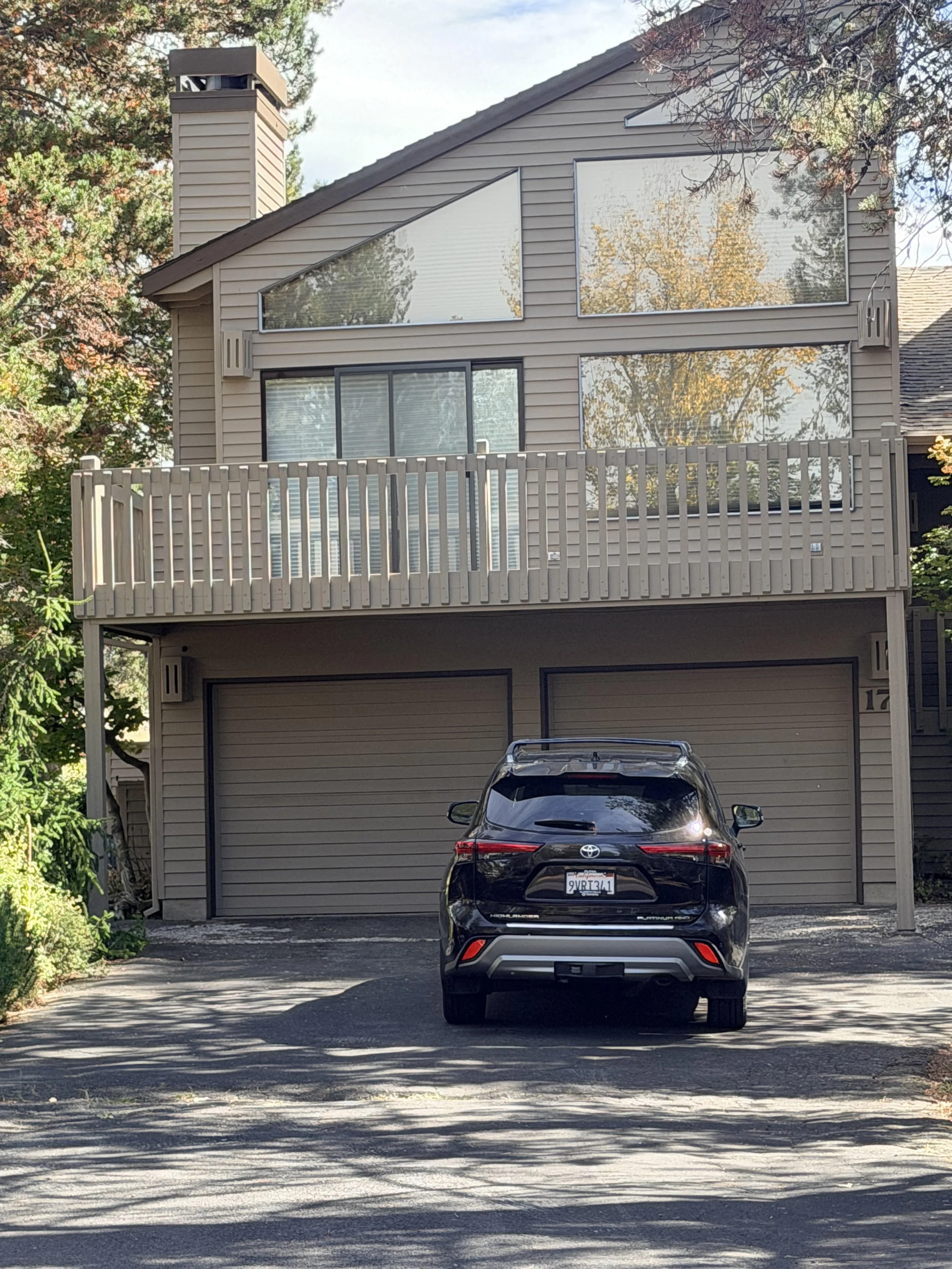 A black Toyota SUV parked in front of a two-story house with a tan wooden exterior and a garage door. The house has large windows on the second floor and a garage on the ground level. There is a driveway with some shadows from trees, and part of a tr