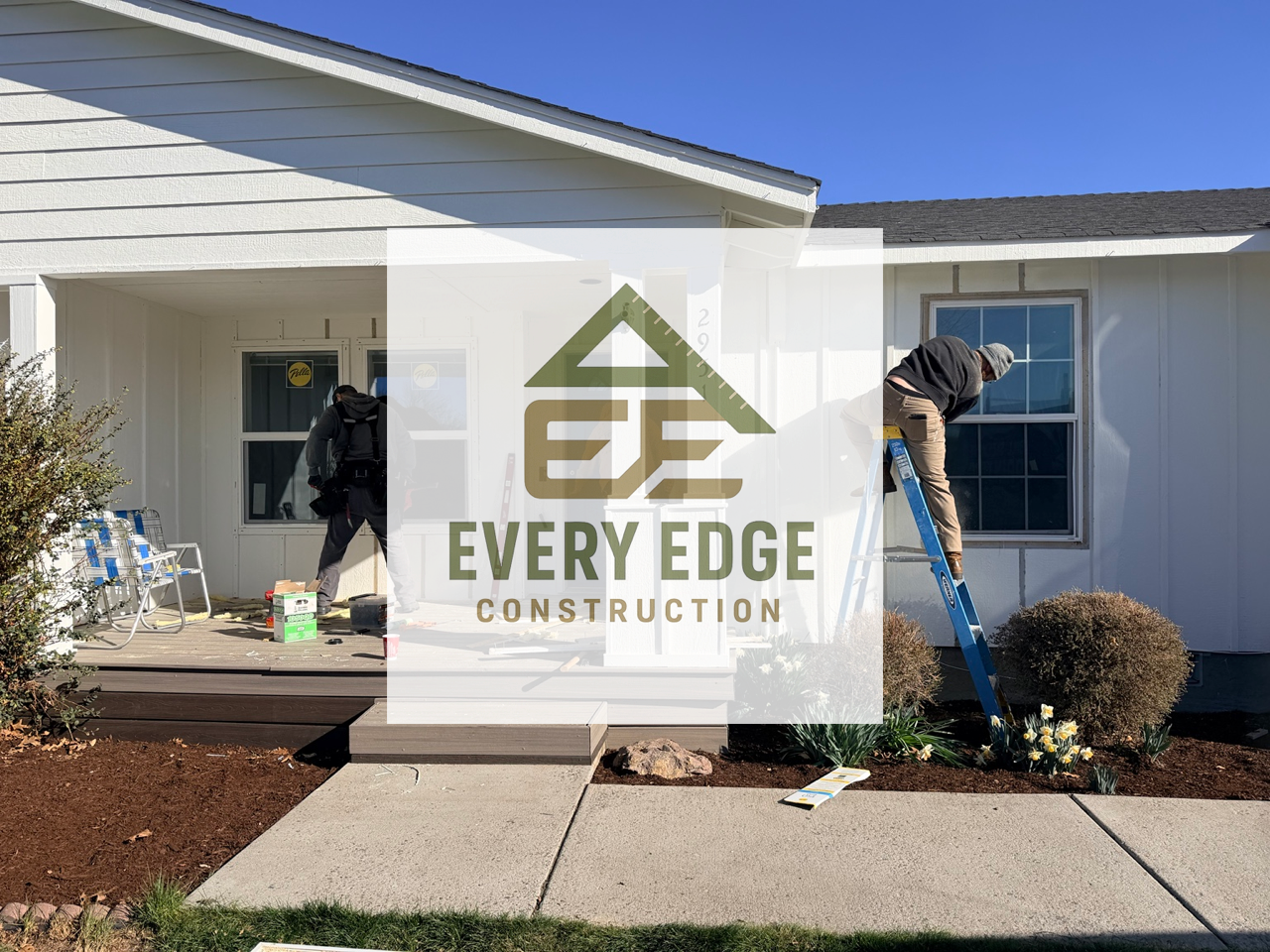 Construction workers working on the exterior of a white house with a porch and window, landscaping with bushes and flowers, and a clear blue sky.