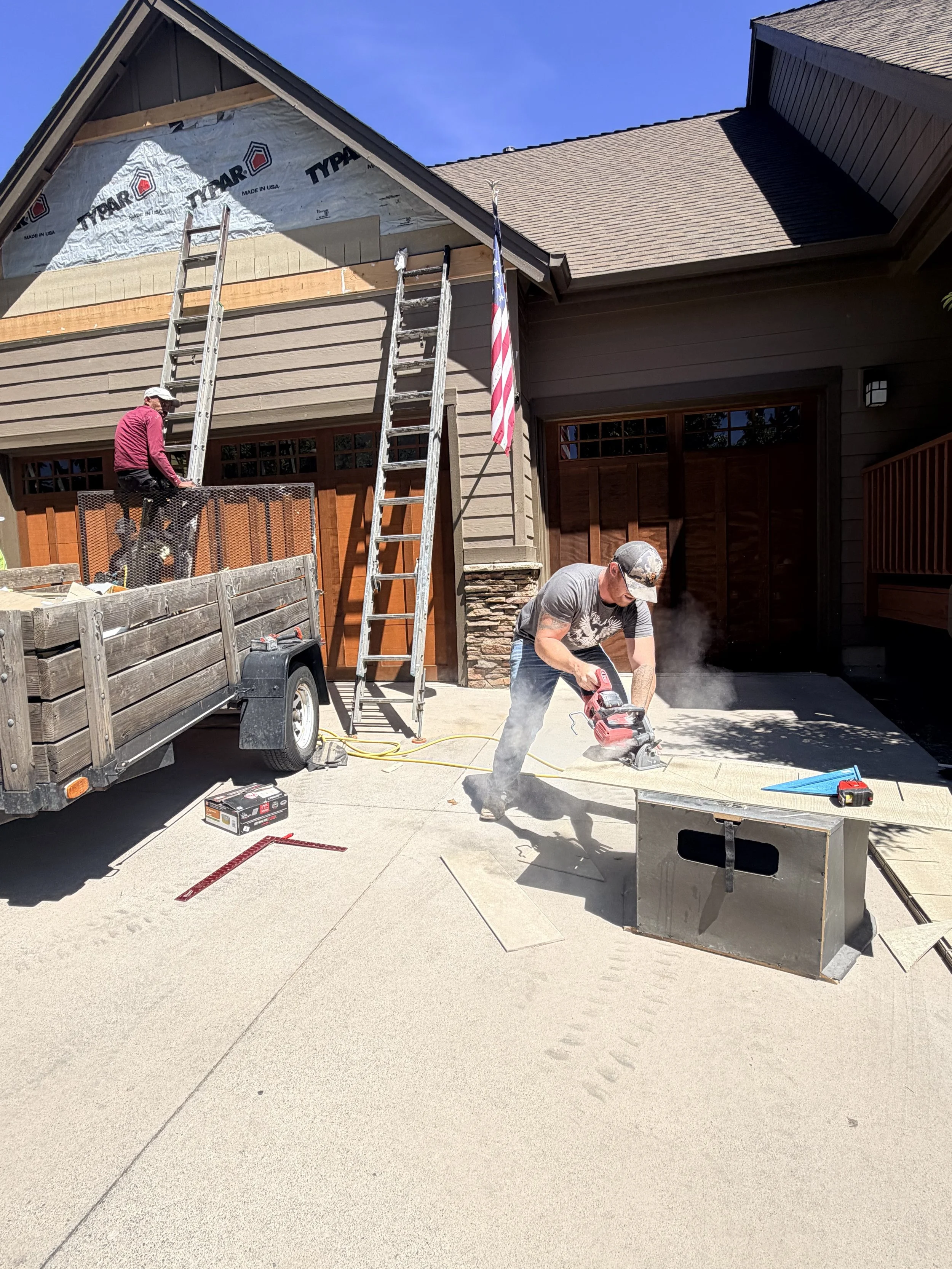 Two men working on construction or renovation of a house in a driveway, with ladders and tools, one cutting material on the ground and the other on a ladder near the house.