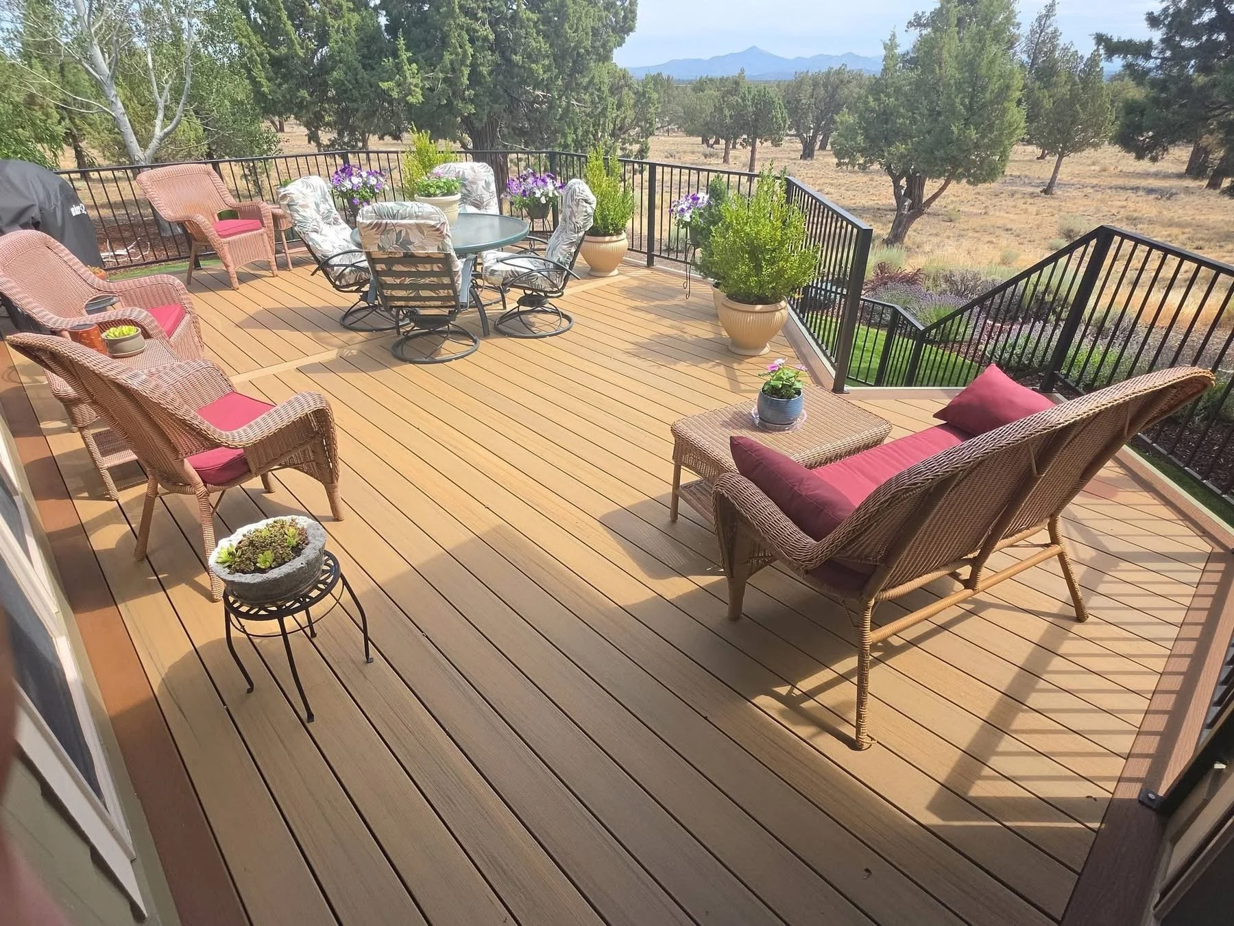A wooden patio deck with outdoor furniture, including wicker chairs with red cushions, a glass-top table, and metal chairs with patterned cushions, surrounded by potted plants and overlooking a landscape with trees and mountains in the distance.