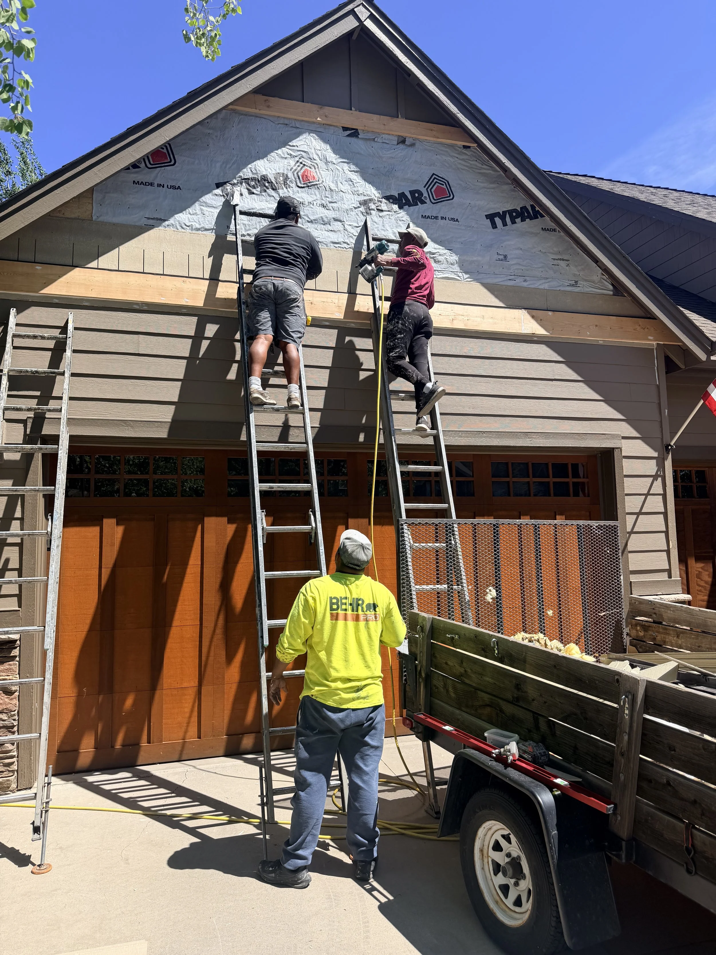 Two workers standing on ladders installing house siding, with a man in a yellow safety jacket observing and a truck carrying materials parked beside a beige house under a clear blue sky.