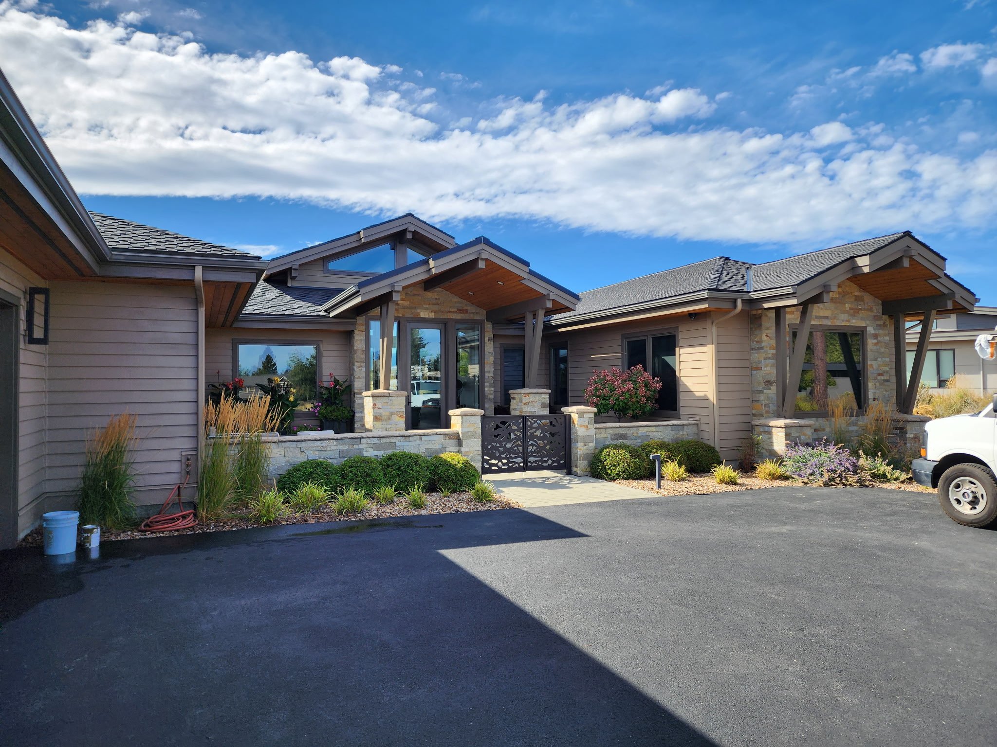 A modern house with a gray shingled roof, large windows, and a small garden, under a partly cloudy blue sky.