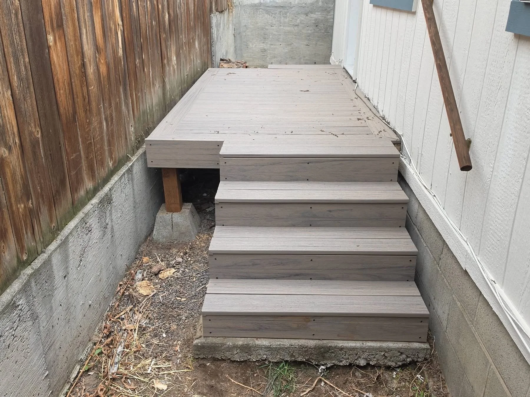 A small outdoor staircase made of light-colored wood attached to the side of a house, with a wooden handrail on the right side, surrounded by a concrete foundation and an unpaved ground area with dirt and some dried leaves.