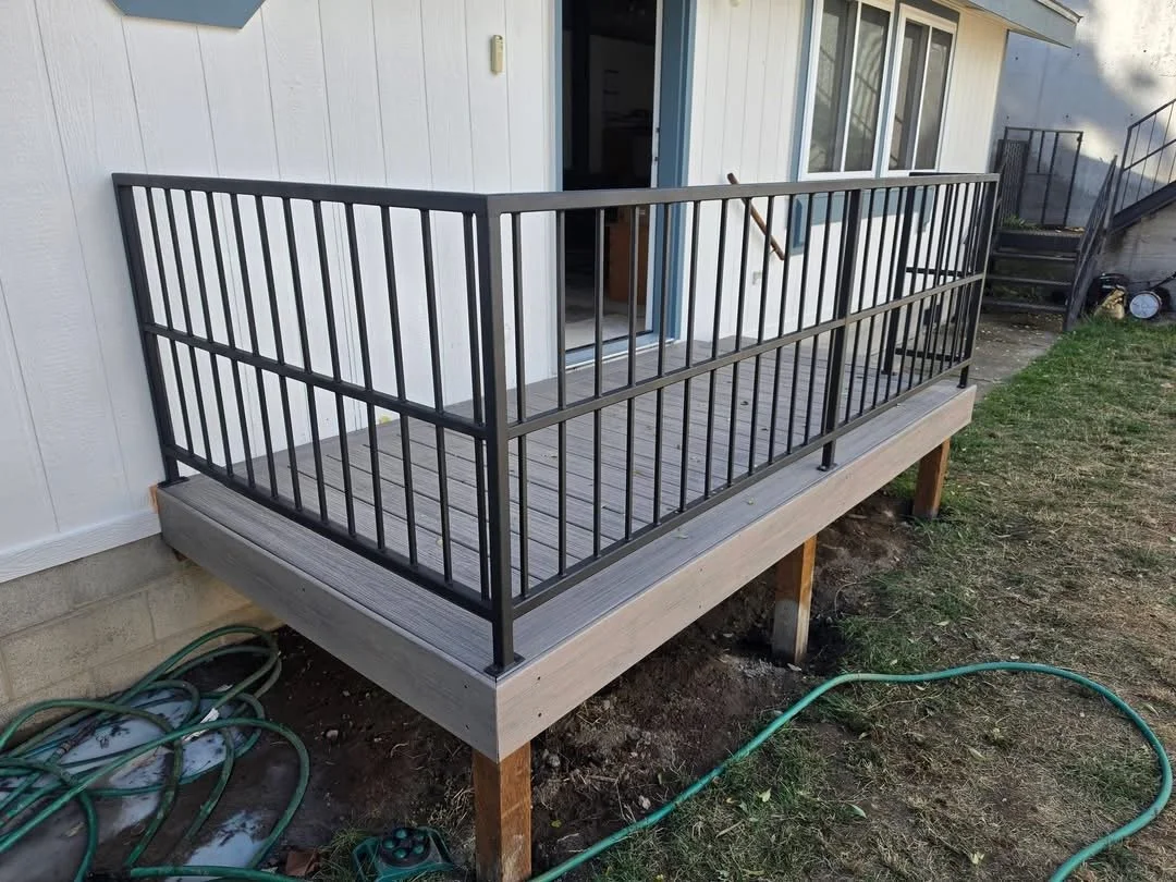 Wooden deck with black metal railing attached to the back of a white house, with a sliding door leading inside, and outdoor stairs on the side.
