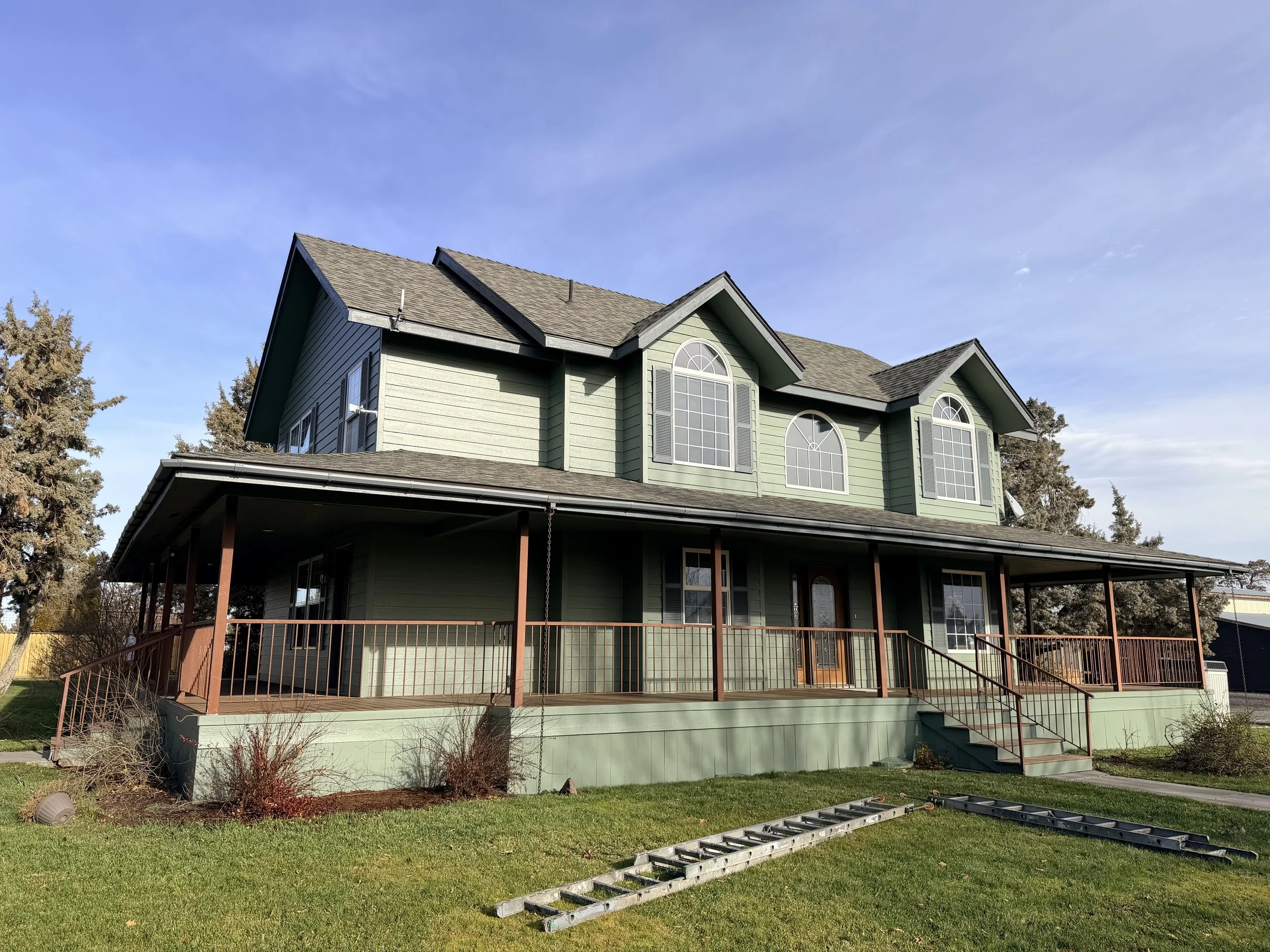 A two-story green house with large arched windows, a wooden deck, and a set of gray ladders on the grass in front, under a blue sky with some clouds.