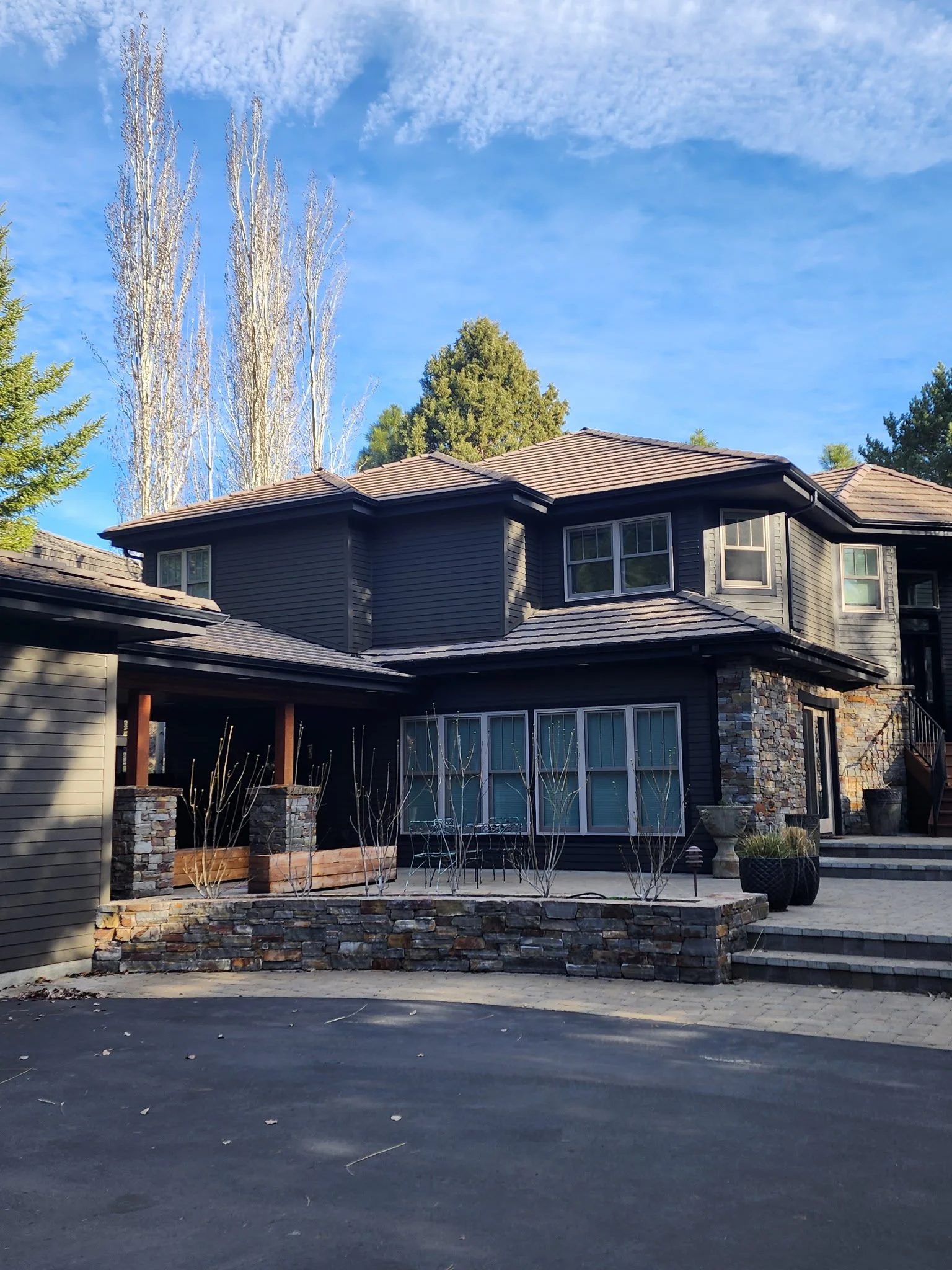 A modern two-story house with a gray exterior, stone accents, multiple windows, and a tiled roof. There is a paved driveway in front and small landscaping elements including trees and planters.