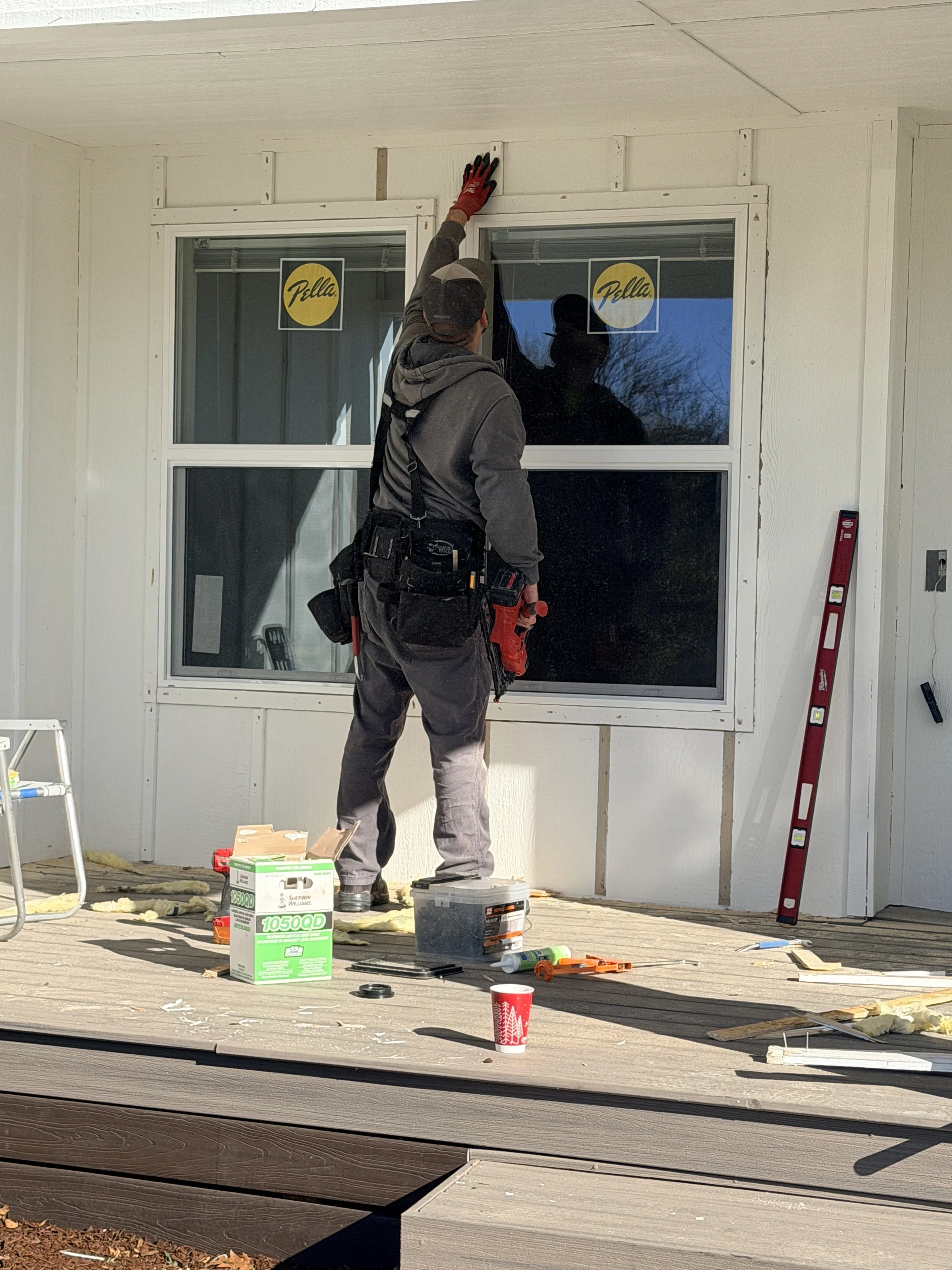 A construction worker installing or repairing a window on a house exterior. The worker is reaching up to the ceiling, wearing work gloves, a hoodie, and a tool belt. There are construction materials and tools scattered on the deck below, including a level, a box, and a cup.