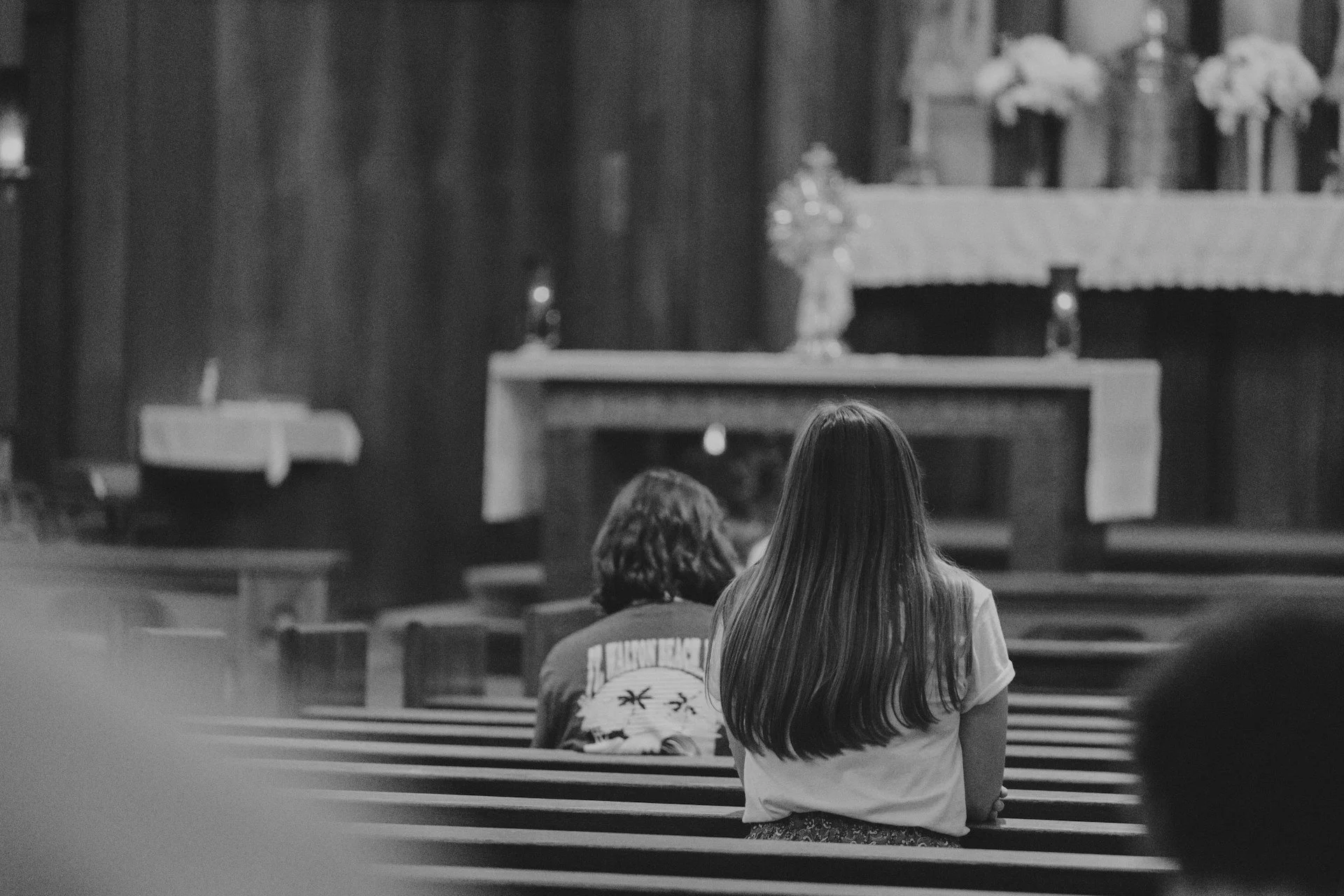 Two women sitting in pews at the back of a church during a service, facing the altar decorated with flowers and religious items, in black and white.