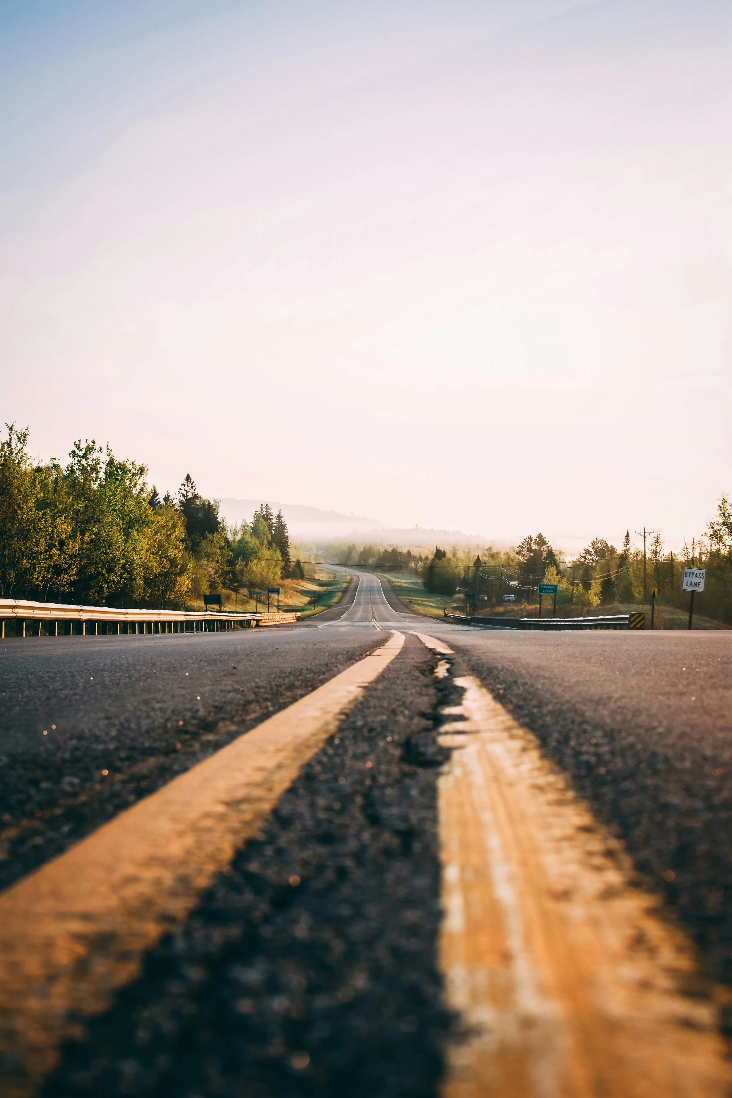 A scenic rural road at sunrise or sunset, with double yellow lines in the foreground, trees on both sides, and a misty horizon in the distance.