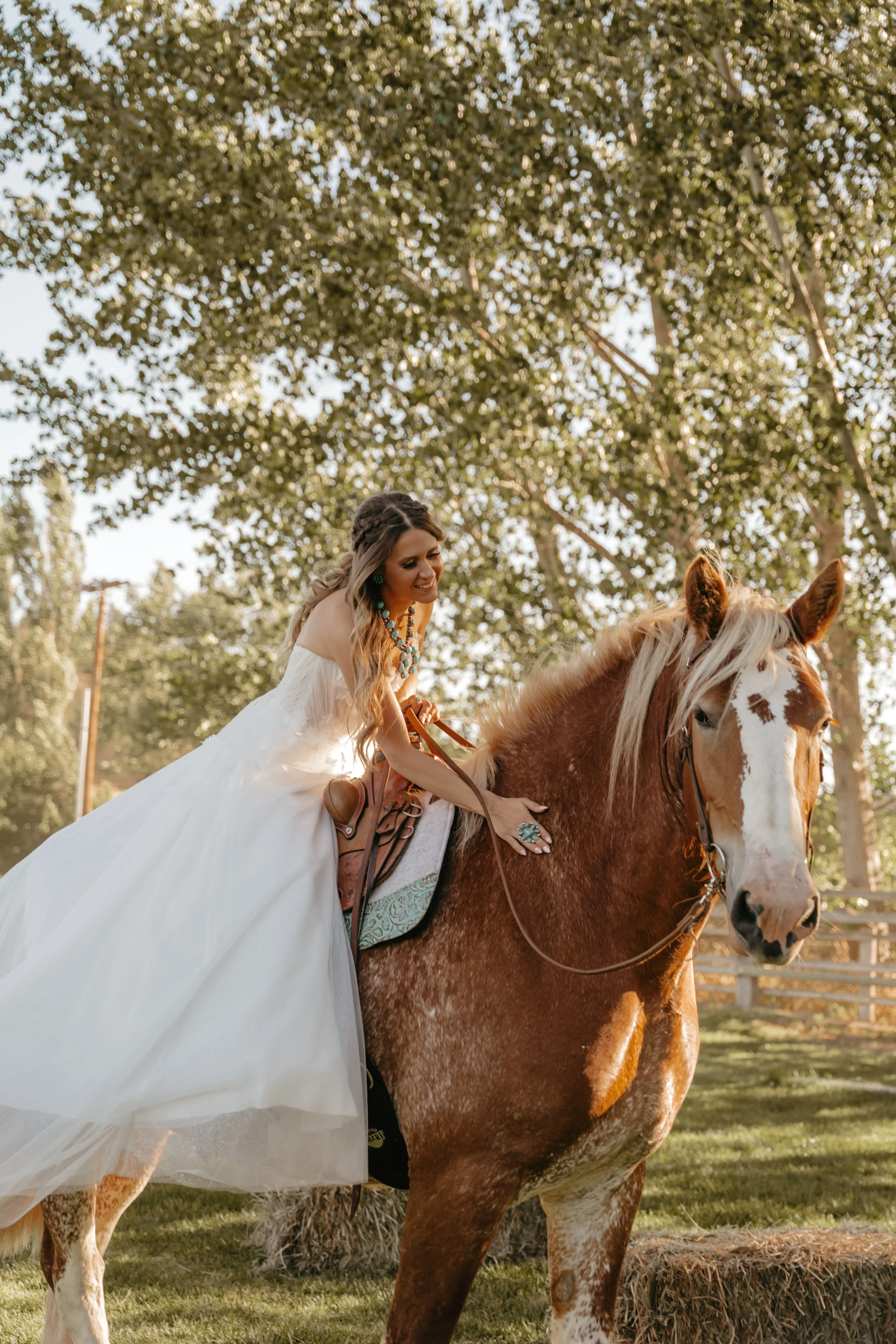 A woman in a white dress riding a brown and white horse outdoors. She is smiling, wearing jewelry, and surrounded by trees and sunlight.