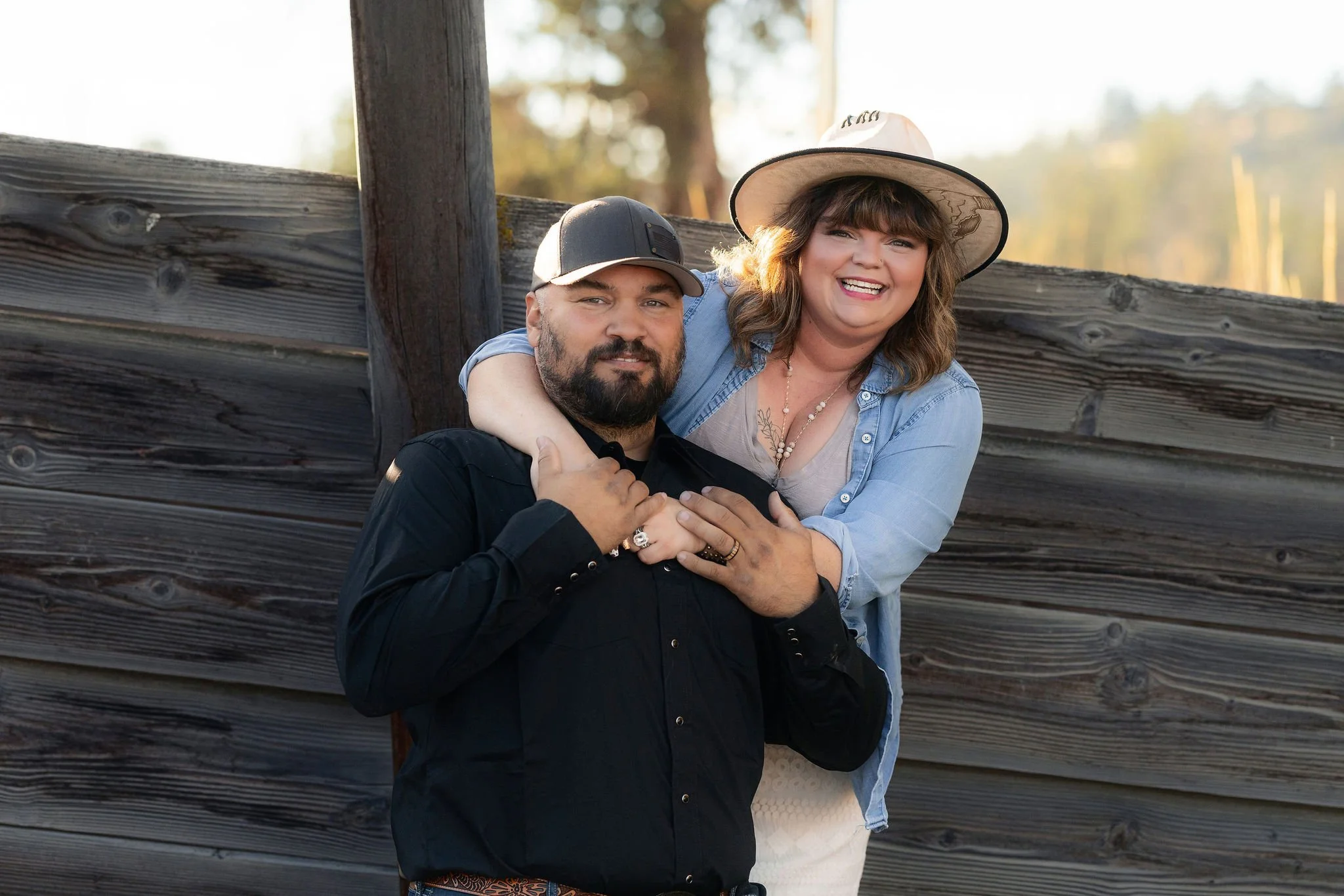 A smiling woman wearing a wide-brimmed hat, denim shirt, and necklace, and a man wearing a baseball cap and dark shirt, posing together outdoors near a wooden fence.