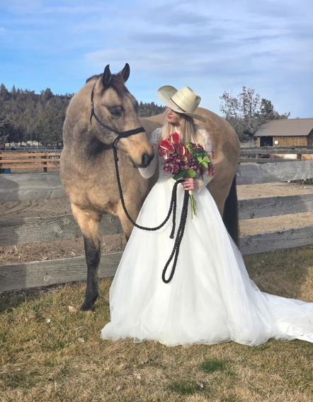 A woman in a white wedding dress holding a bouquet of flowers standing next to a gray horse at a fenced outdoor farm with trees and a barn in the background.