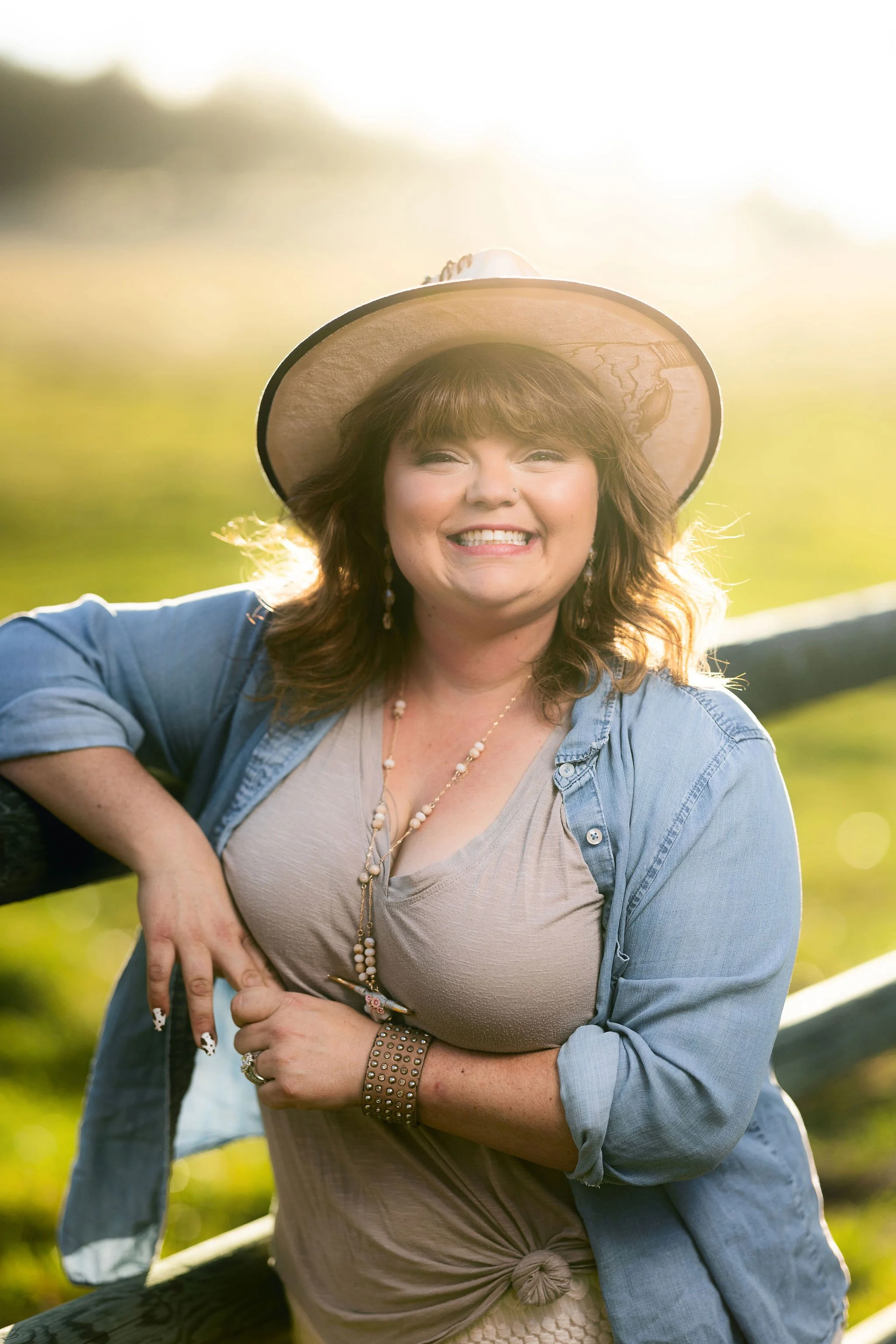 A woman smiling outdoors wearing a wide-brimmed hat, denim jacket, beige top, and jewelry, with greenery and sunlight in the background.