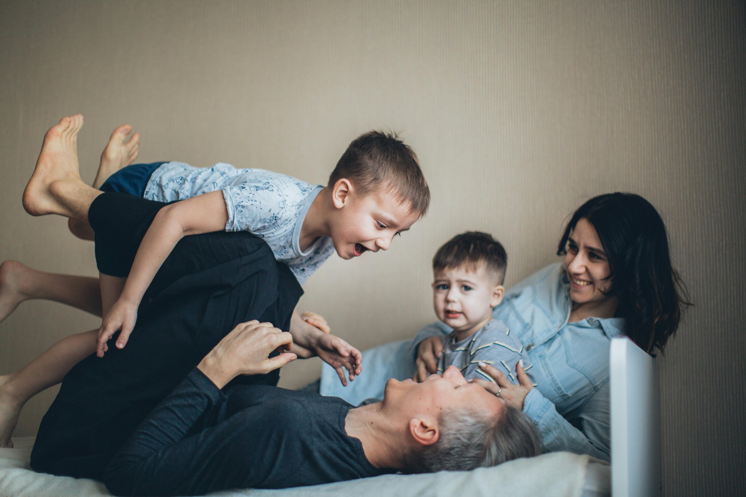 A family of four, including a woman, a man, and two young boys, playing and laughing on a bed in a bedroom.