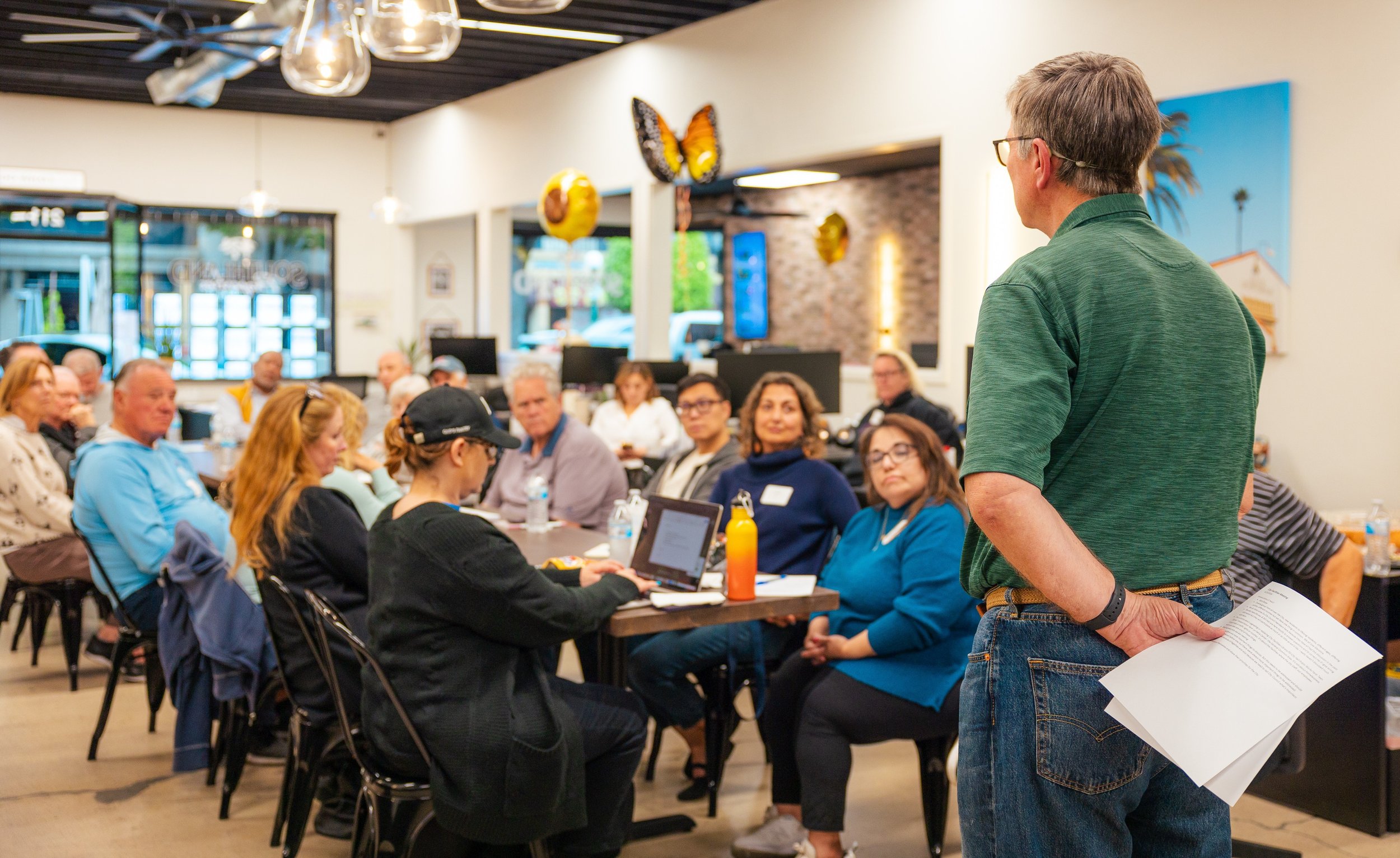 A man in a green shirt and jeans addresses a group of people sitting at tables in a casual restaurant or cafe. The group listens attentively, with some taking notes or looking at their devices, while the man holds papers in his left hand.