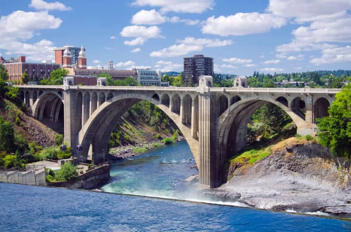 A large concrete arch bridge spanning a river with a dam feature, surrounded by green trees and a city skyline in the background under a partly cloudy sky.