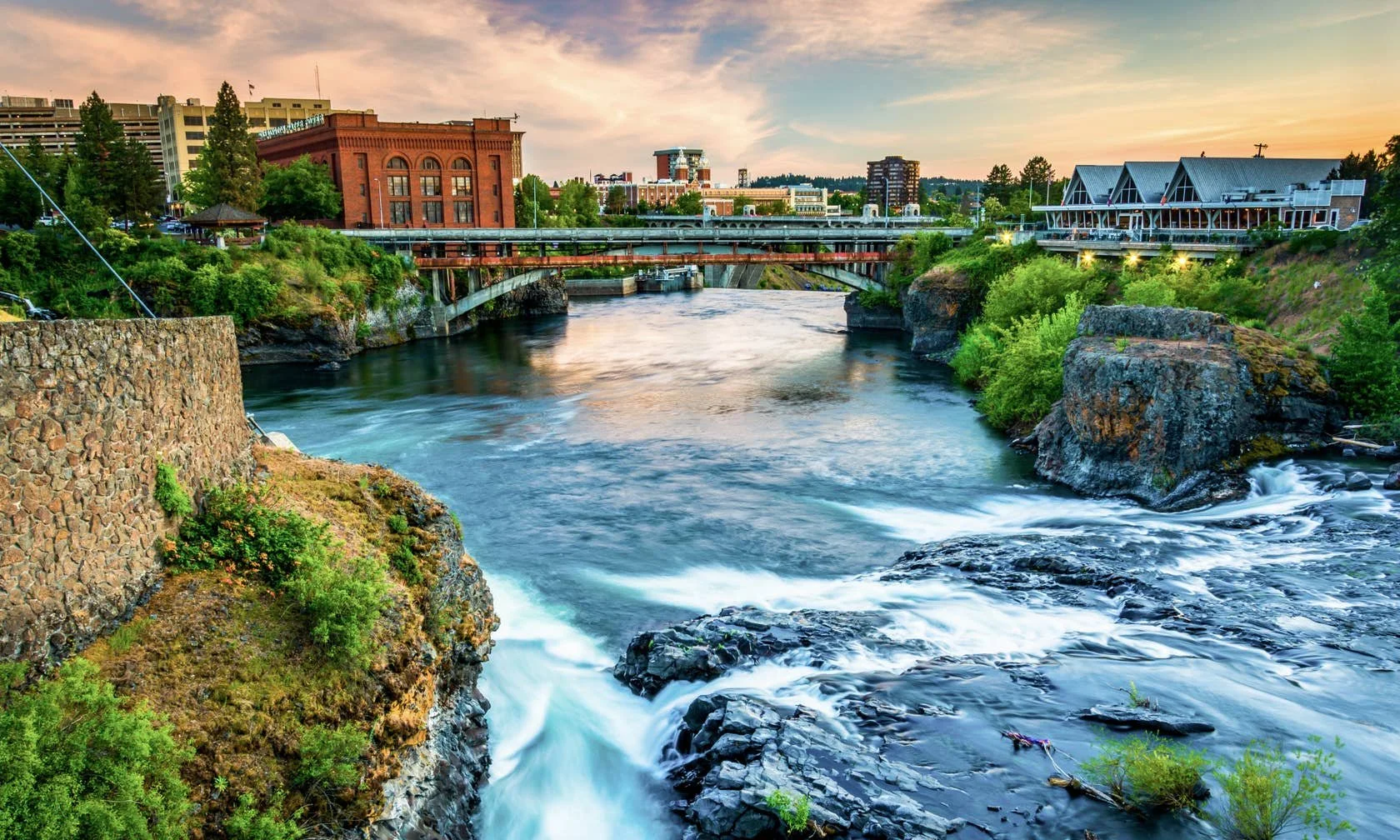 A city river scene at sunset with flowing water, rocks, greenery, and buildings in the background.