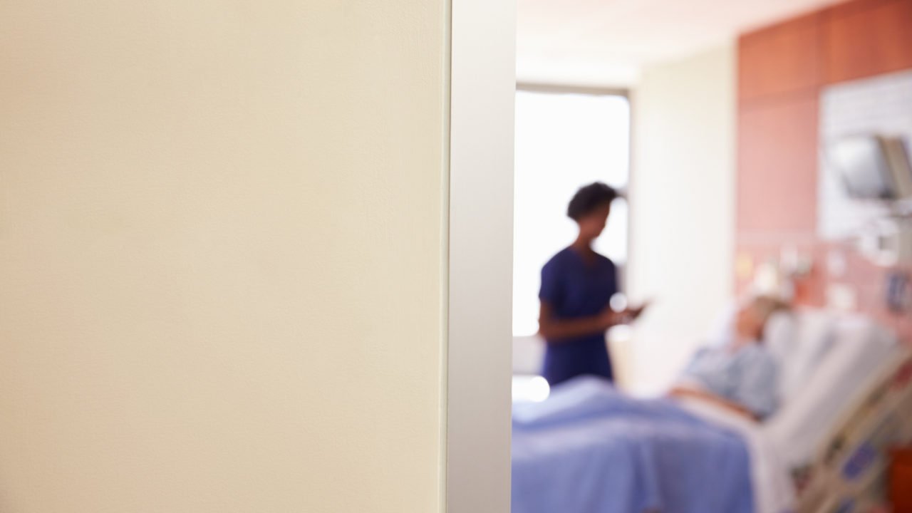 A hospital room with a blurred view of a nurse using a tablet and a patient lying in bed.