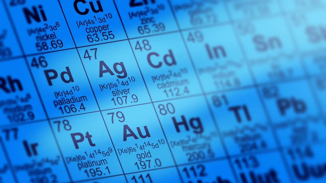 Close-up of a periodic table showing chemical symbols and atomic numbers for elements like Nickel, Copper, Silver, Gold, and Platinum, with a blue overlay.