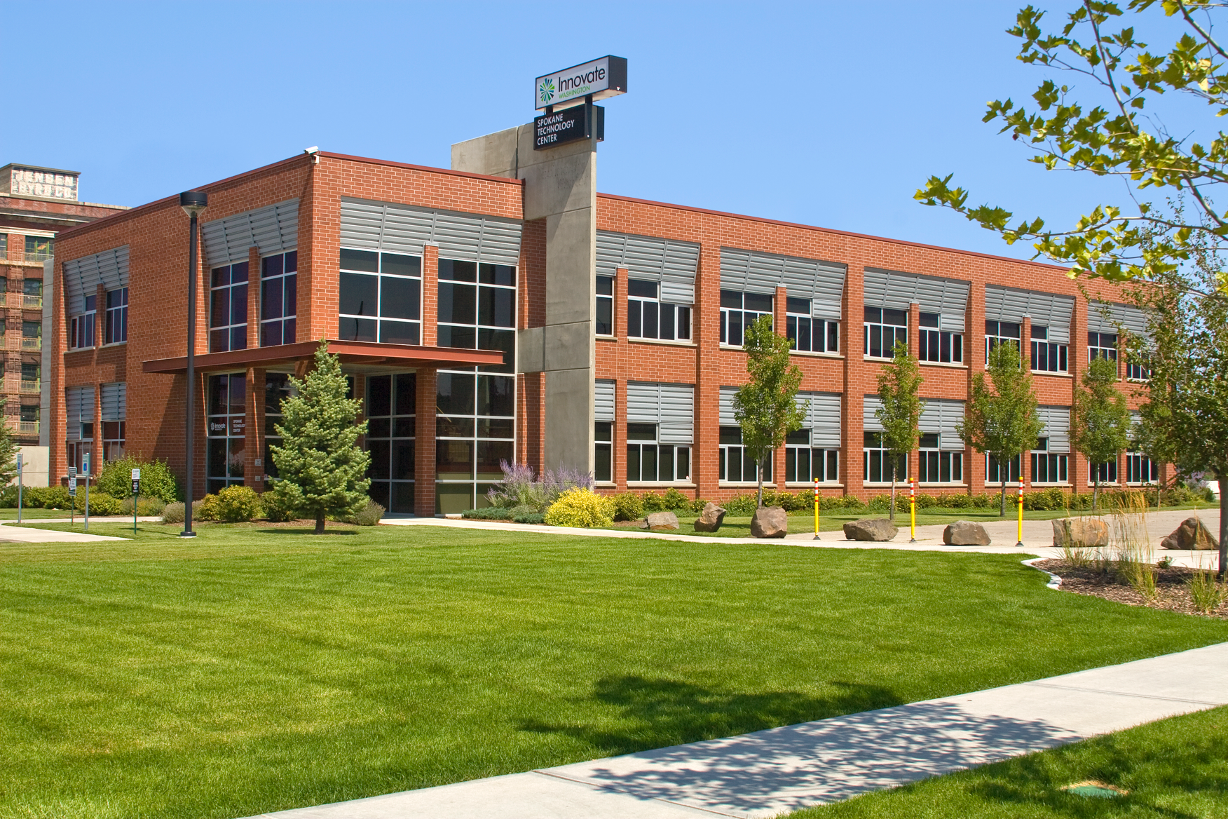 Modern red brick building with large windows and a sign for the Spokane Technology Center at Innovate Washington, surrounded by well-maintained grass and trees.
