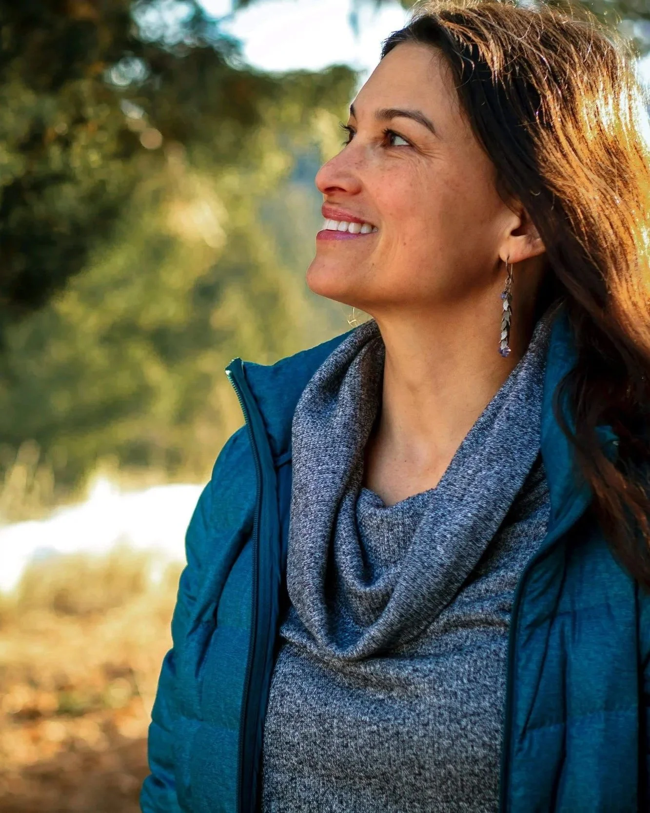 A smiling woman with long, dark hair outdoors, wearing a blue jacket, gray sweater, and earrings, with trees and sunlight in the background.