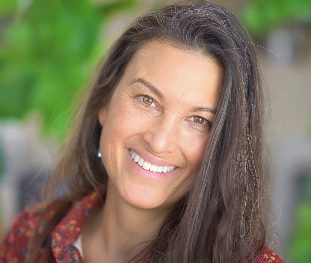 Close-up of a woman with long brown hair, smiling, with a blurred green outdoor background.