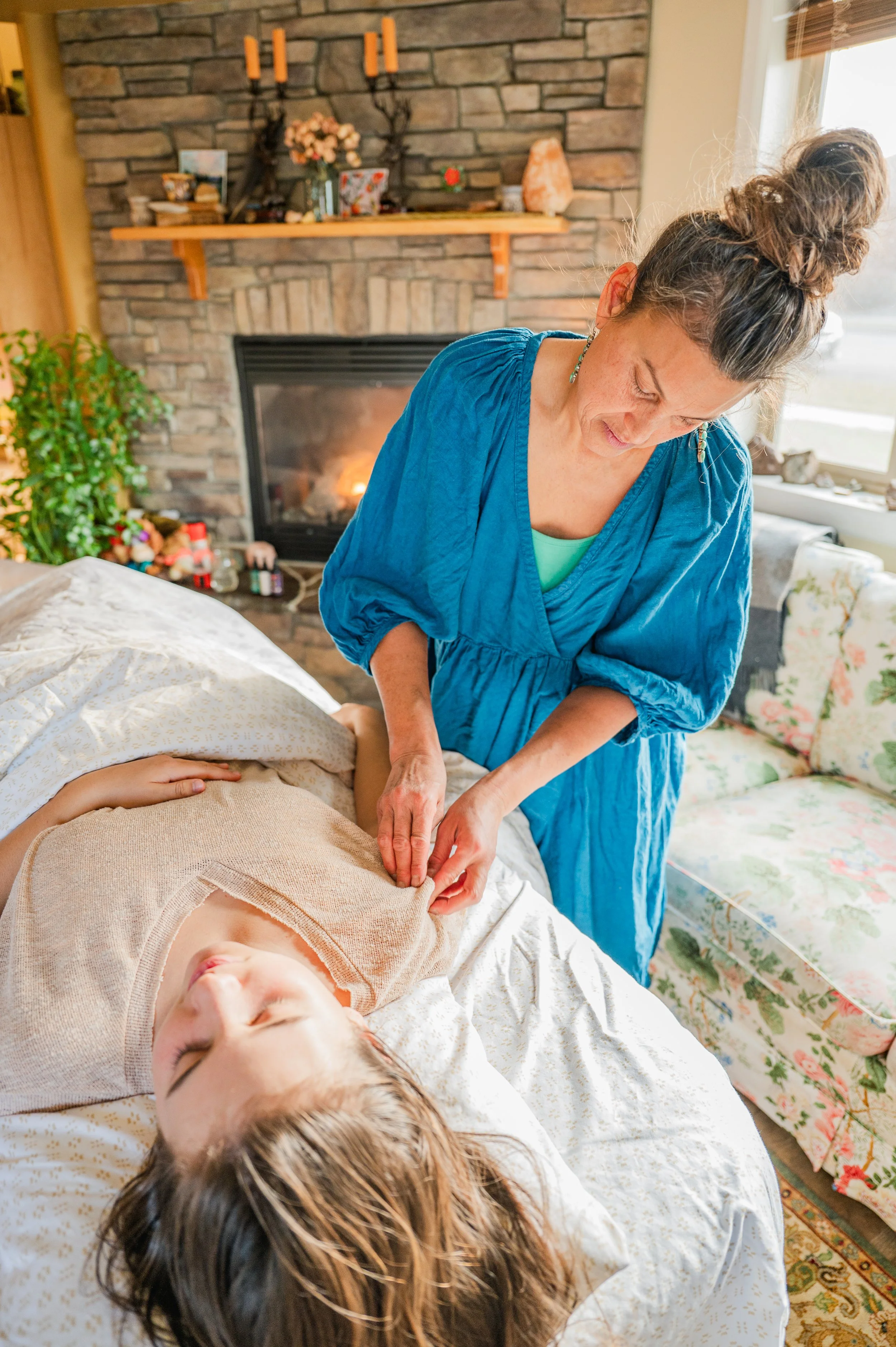 A woman receives a massage from a massage therapist in a cozy living room with a fireplace, bookshelf, and floral couch.