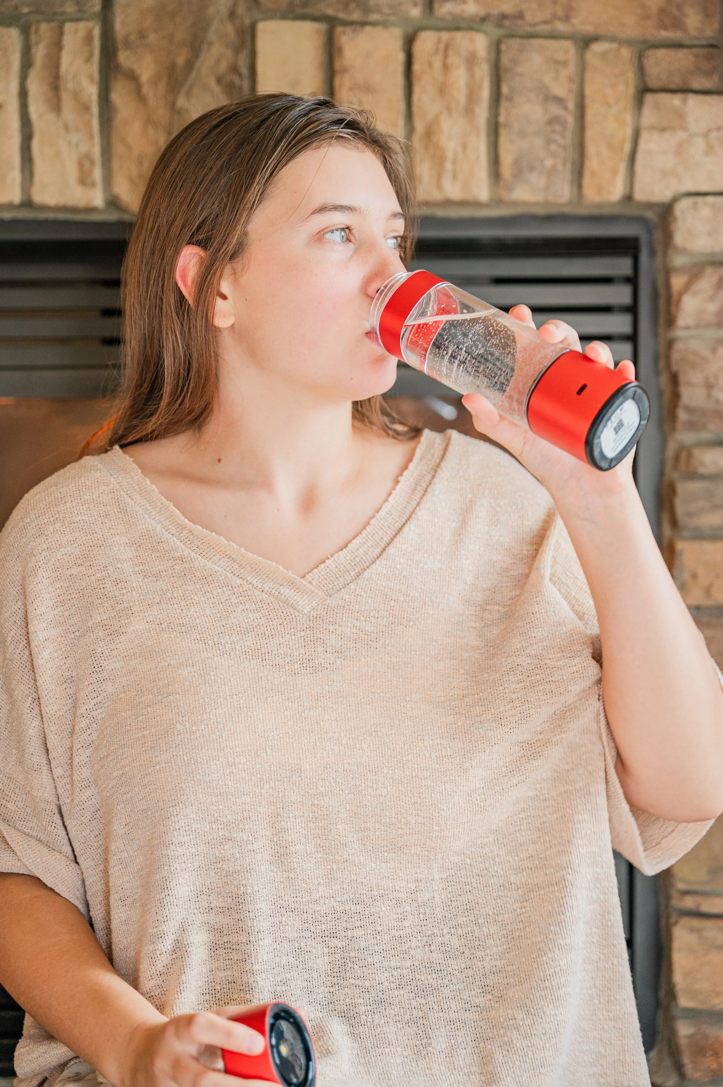 A young woman with brown hair drinking from a clear water bottle with a red lid and holding another similar bottle in her hand.