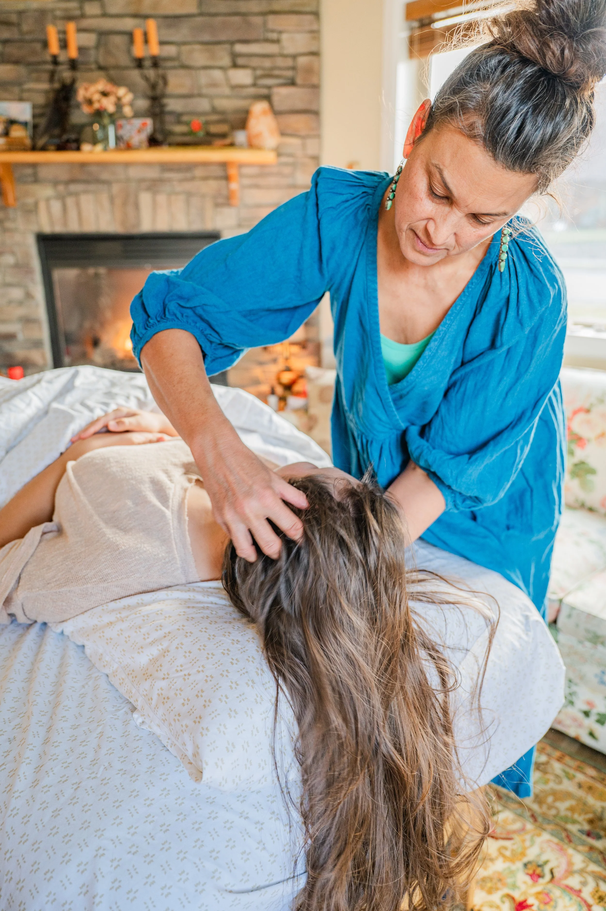 Woman giving a neck massage to a person lying on a bed in a cozy living room with a brick fireplace, decorated with candles and flowers.