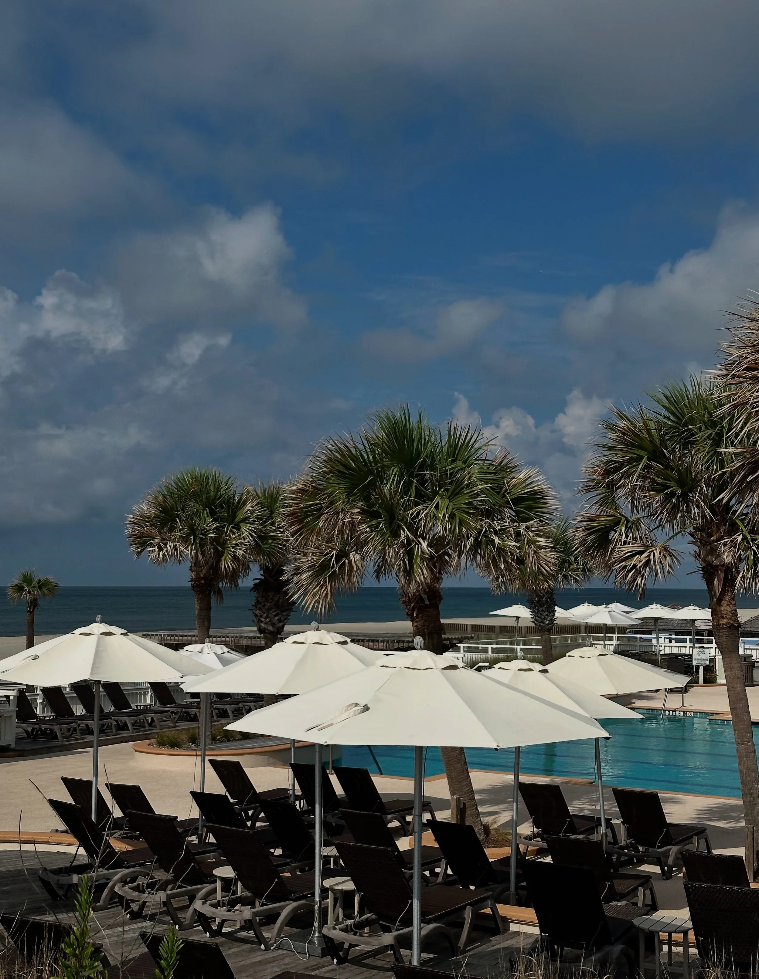 Beachside pool area with lounge chairs, white umbrellas, palm trees, and ocean in the background under a partly cloudy sky.