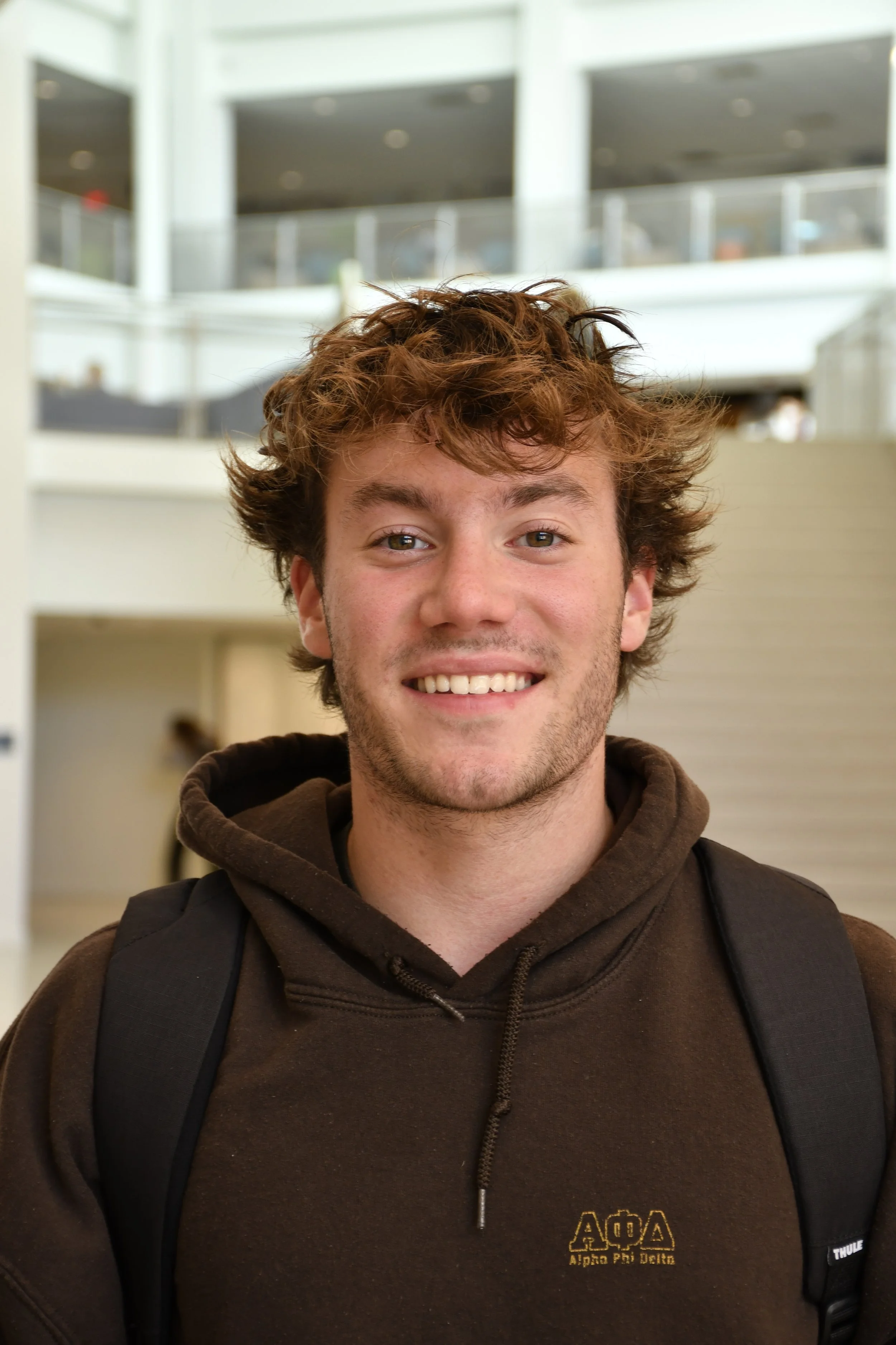 A young man with curly brown hair smiling at the camera, wearing a brown hoodie with Greek letters and a backpack inside a building.