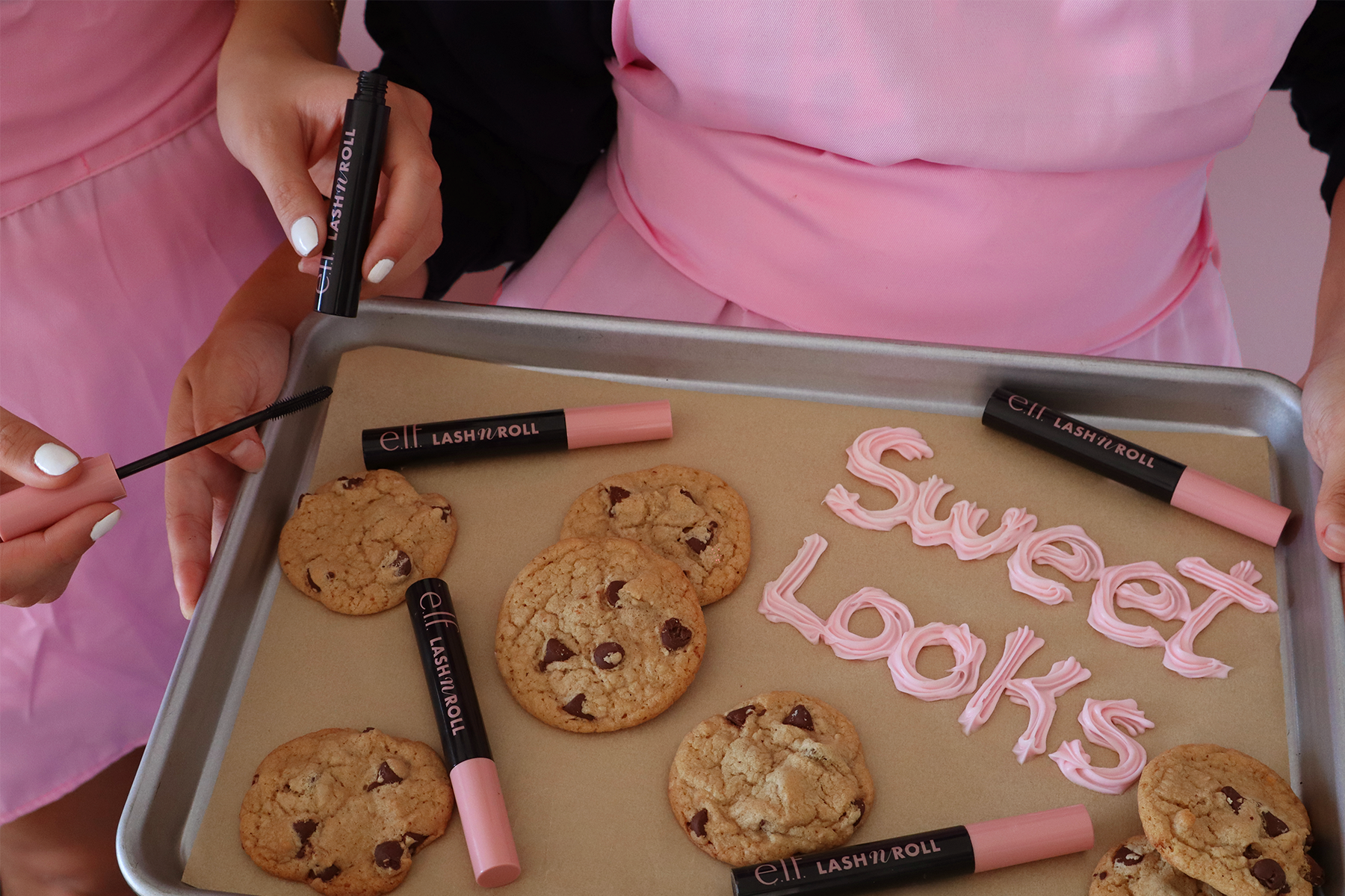 Person decorating cookies with pink frosting spelling 'Sweet Looks' on baking sheet.