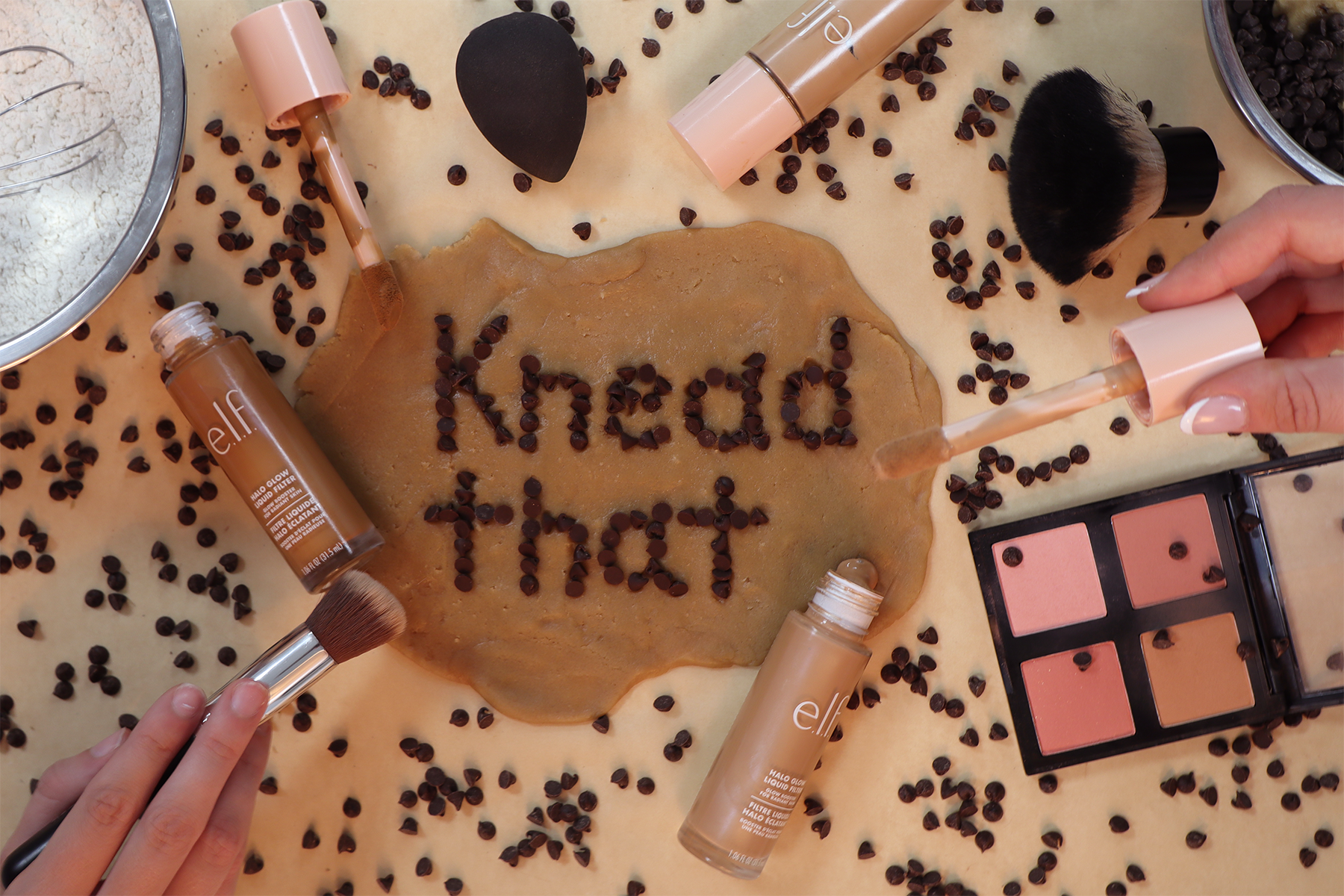 Flat lay of makeup products and chocolate chips on a beige surface with a piece of dough in the center spelling out "Kevin the 1st".