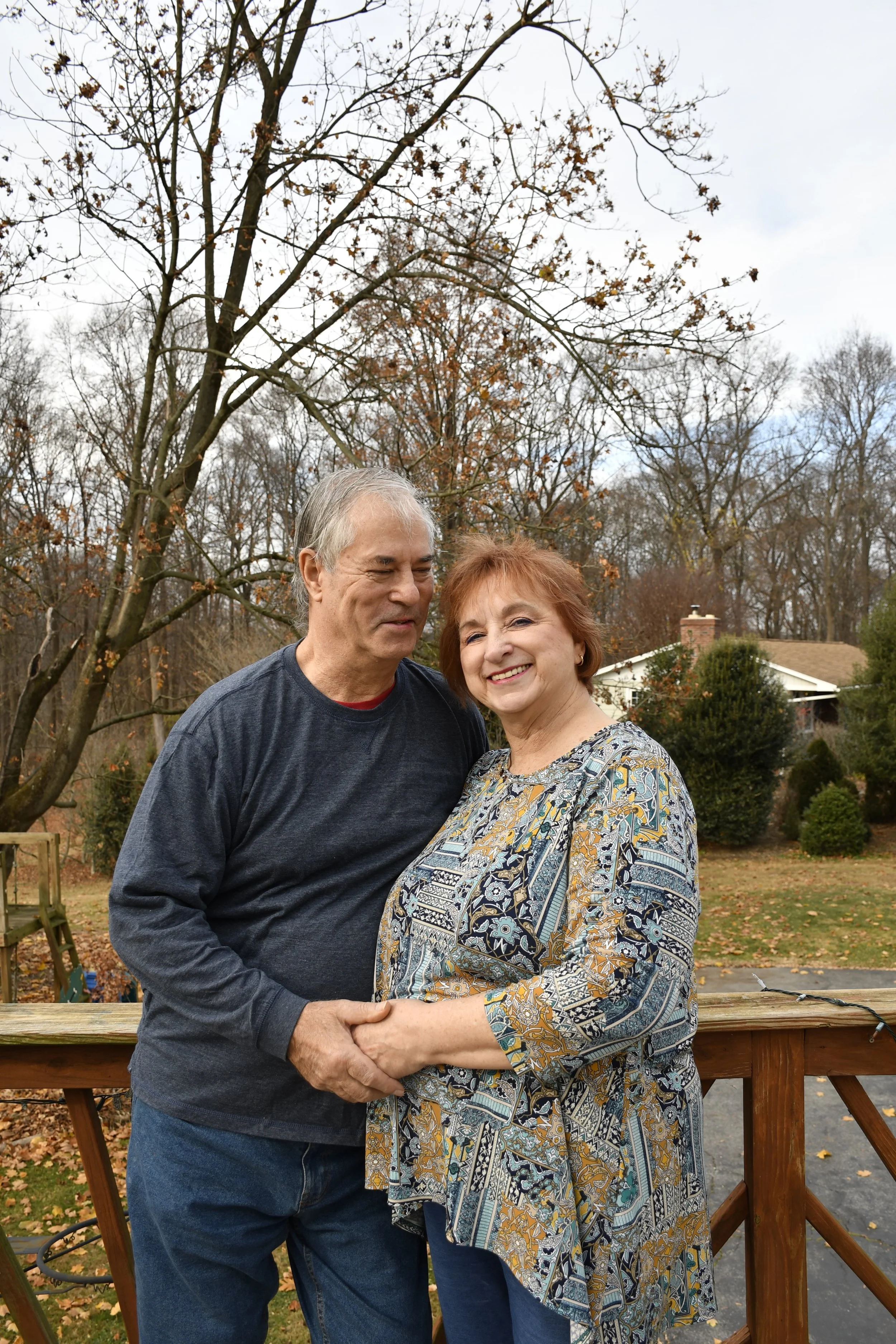 A smiling older woman and a man holding hands on a wooden deck in a backyard with trees and a house in the background during fall.