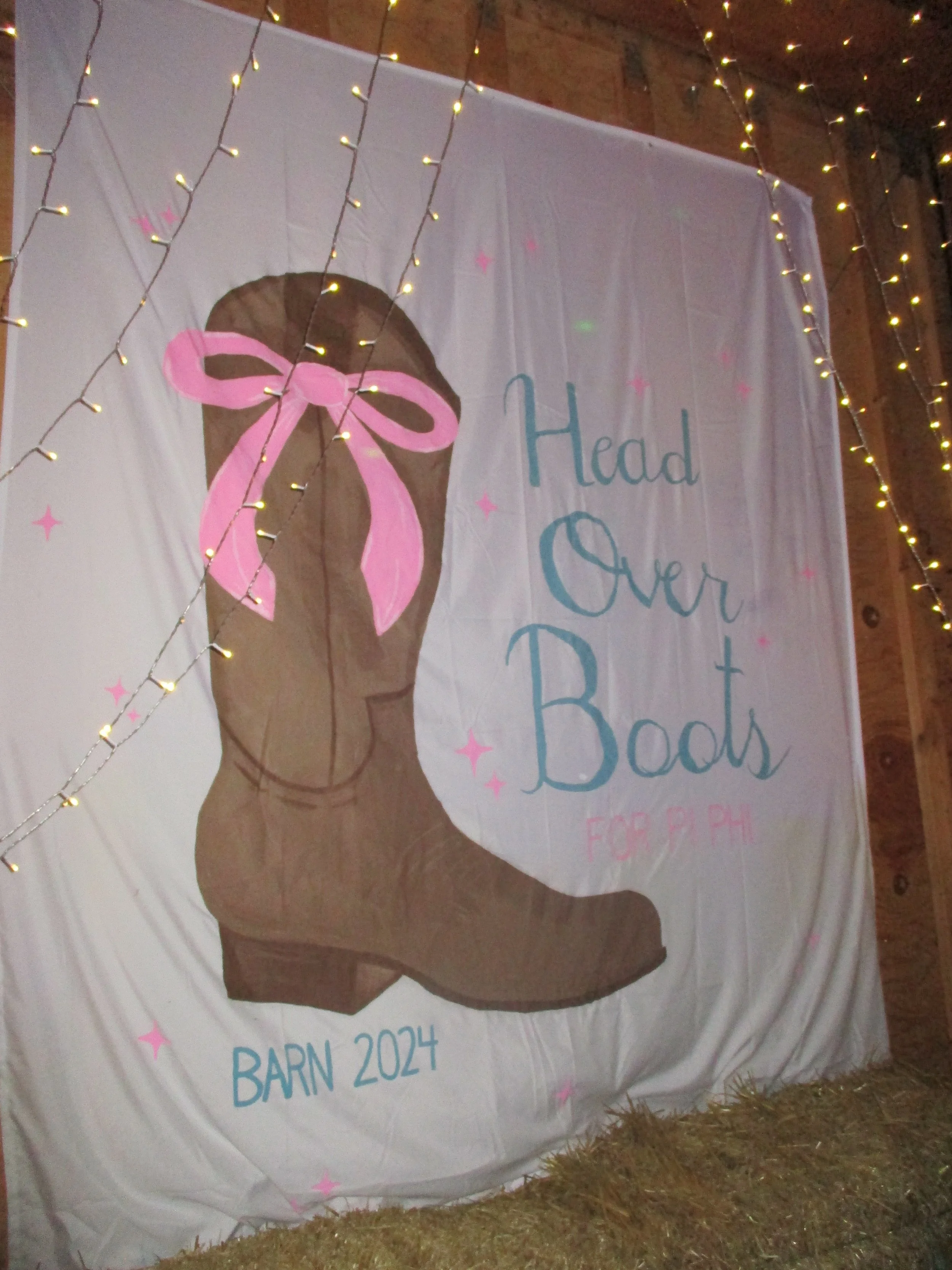 A large fabric banner with a painted image of a brown cowboy boot decorated with a pink bow. The banner contains the text "Head Over Boots for PPH" and "Barn 2024." It is hanging in a barn or rustic setting decorated with string lights and pink star 