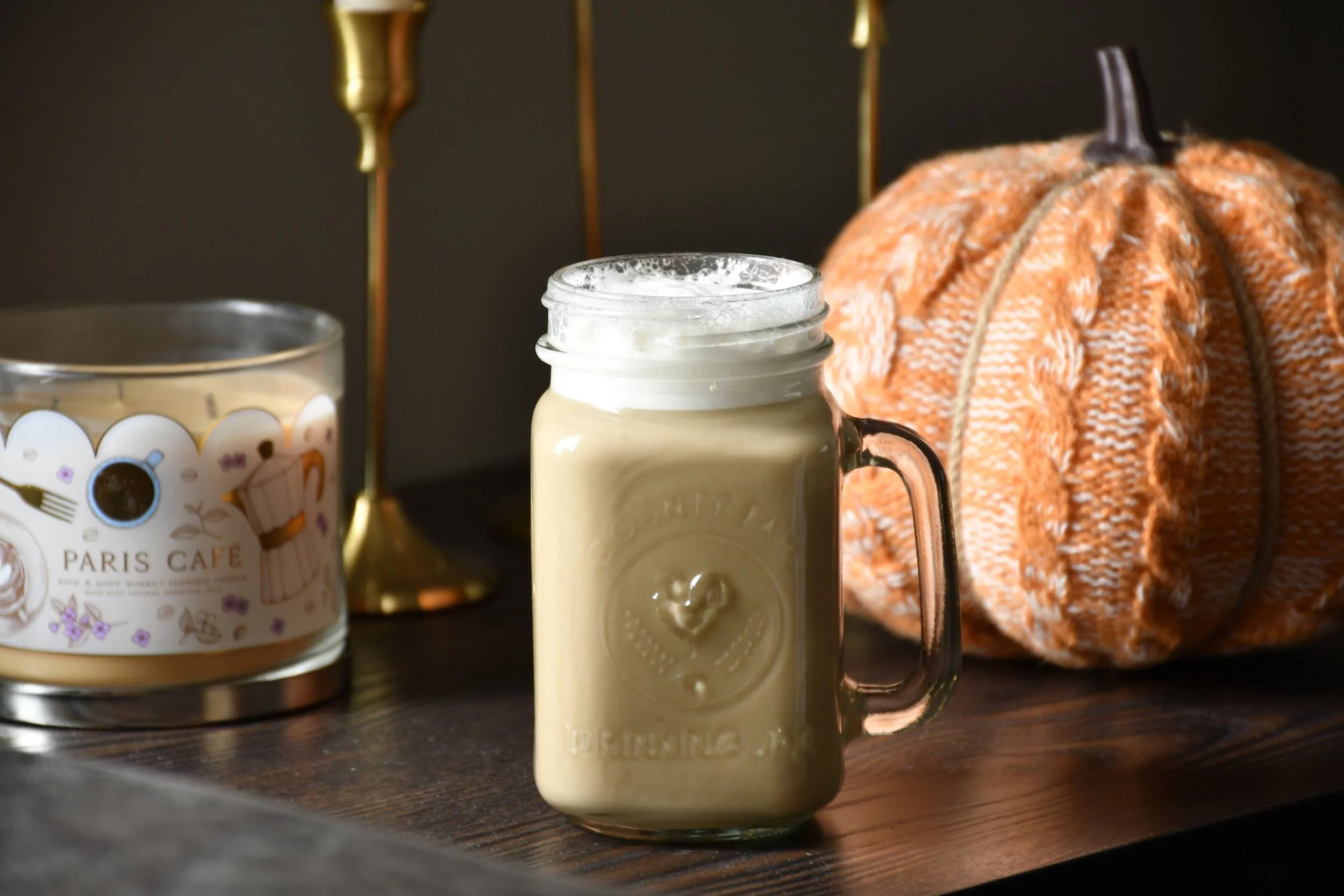 A mason jar filled with a creamy beverage topped with whipped cream, a pumpkin with a textured orange surface, a candle in a glass jar labeled "Paris Café," and a brass lamp stand are arranged on a dark wooden surface.
