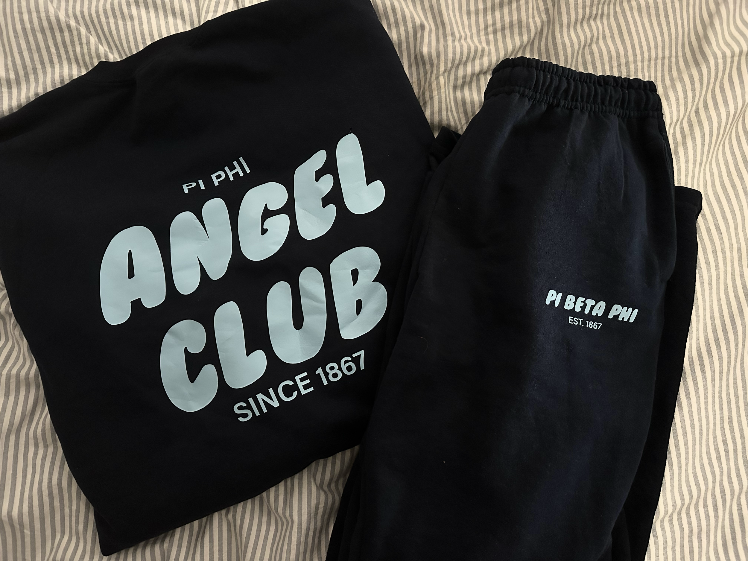 Black sweatshirt and black sweatpants with light blue logos and text from Pi Beta Phi sorority on a striped fabric surface.