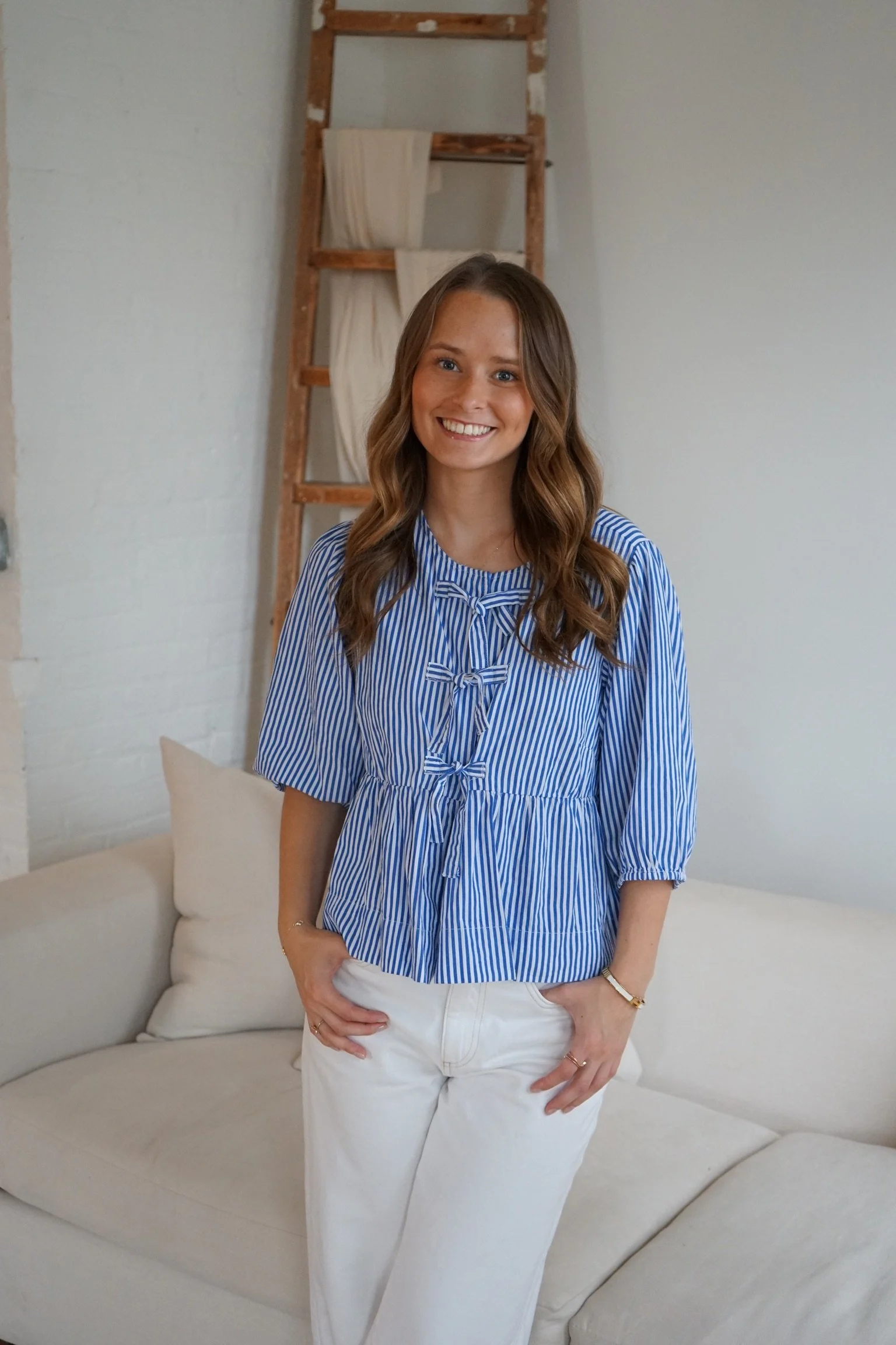 A young woman with long brown hair smiling, wearing a blue and white striped blouse and white pants, standing in a room with a cream-colored sofa and a wooden ladder behind her.