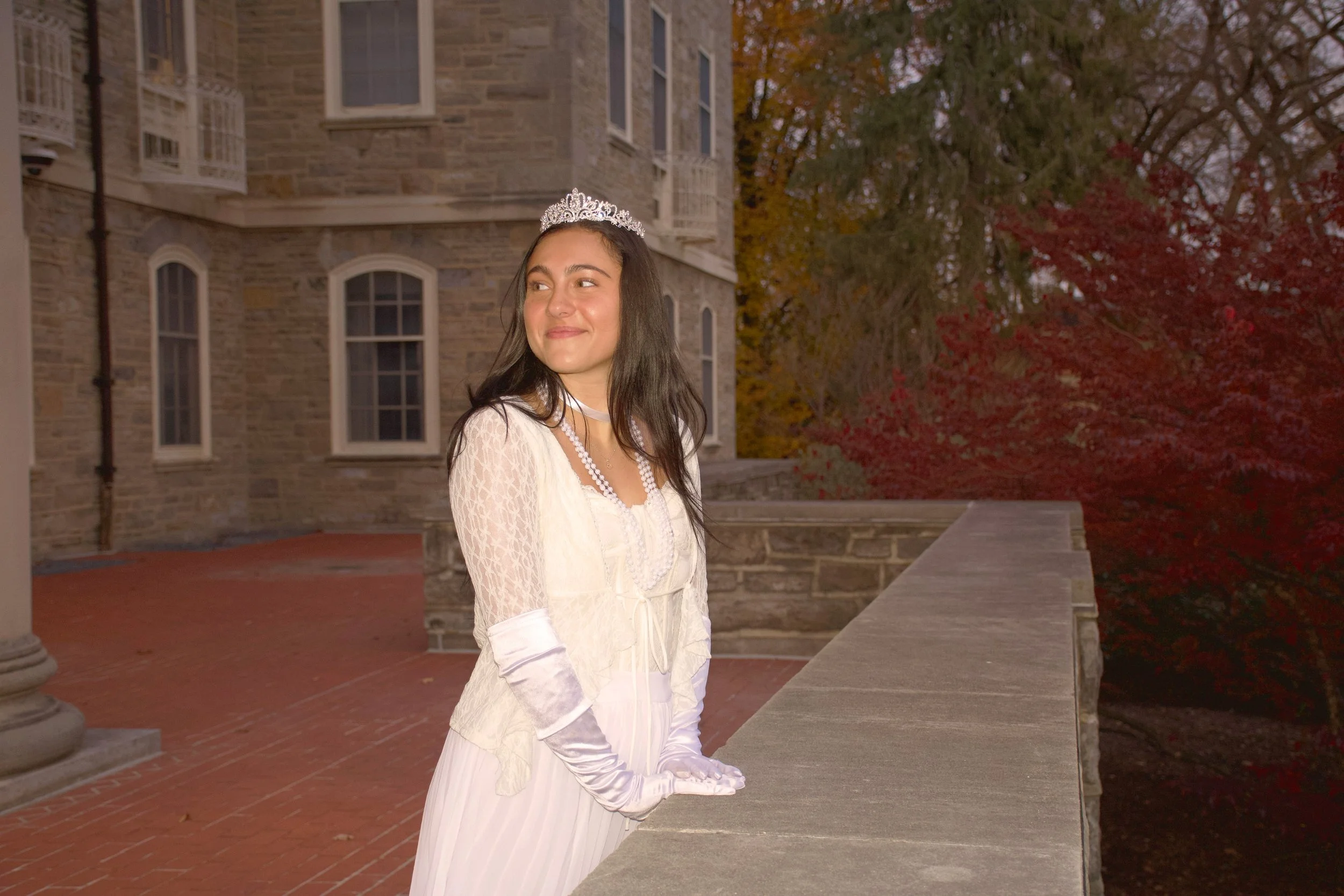 Young woman dressed as a princess in a white gown, lace gloves, and a tiara, standing outdoors on a stone balcony with a historic brick building and autumn trees in the background.