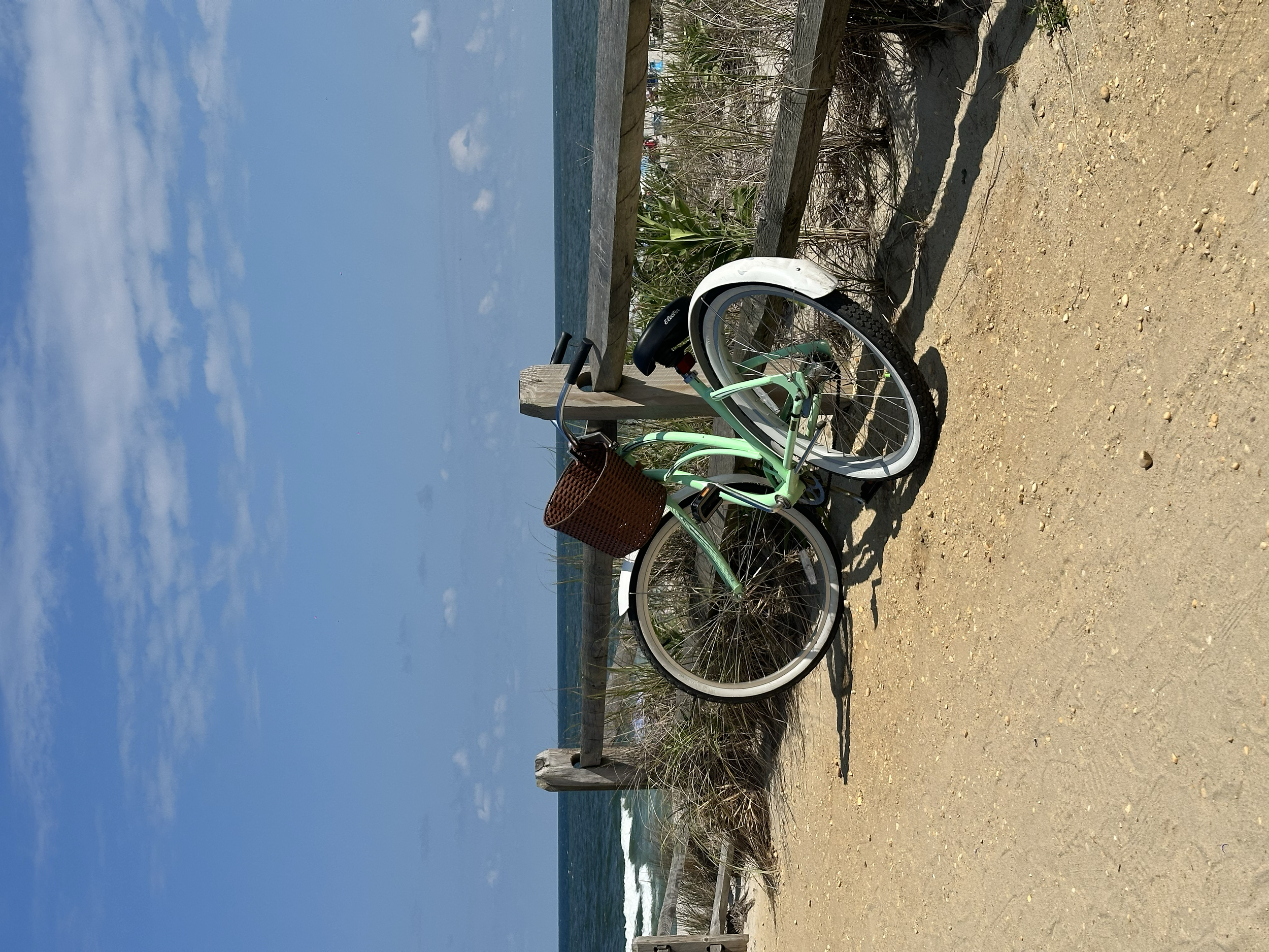 A light green bicycle with a brown woven basket attached to the front, parked against a wooden fence on a sandy beach. The ocean and blue sky with some clouds are visible in the background.